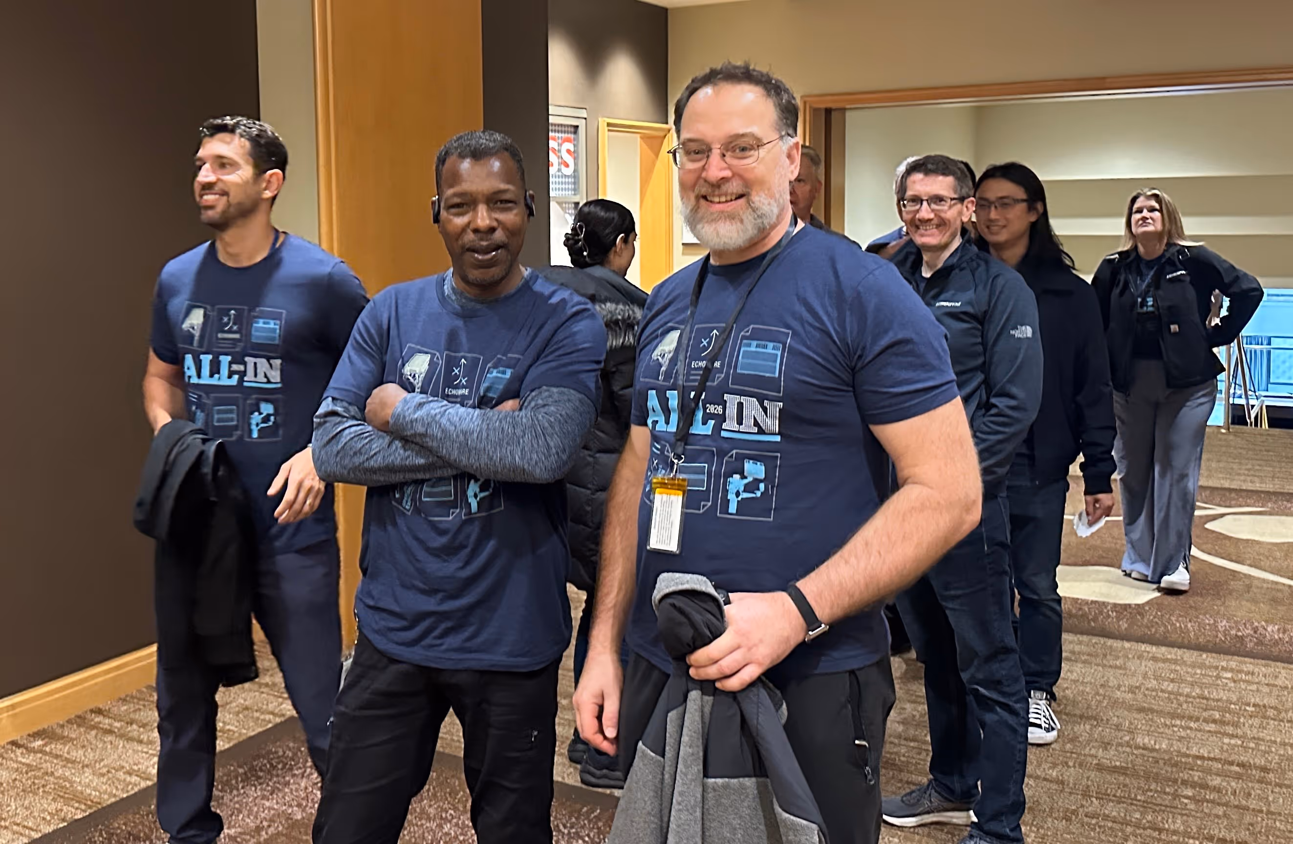 Group of six people indoors, two men in front wearing matching blue All-In T-shirts, others in jackets behind them.