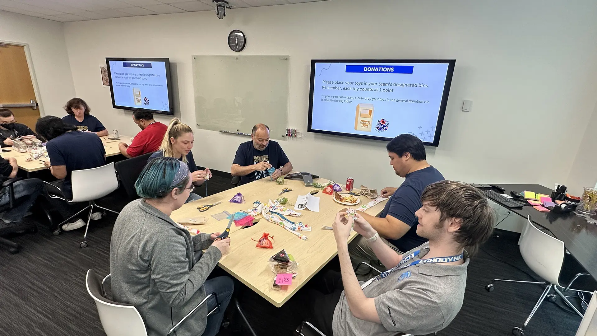 Group of people sitting at tables crafting and reading donation instructions on screens in a meeting room.