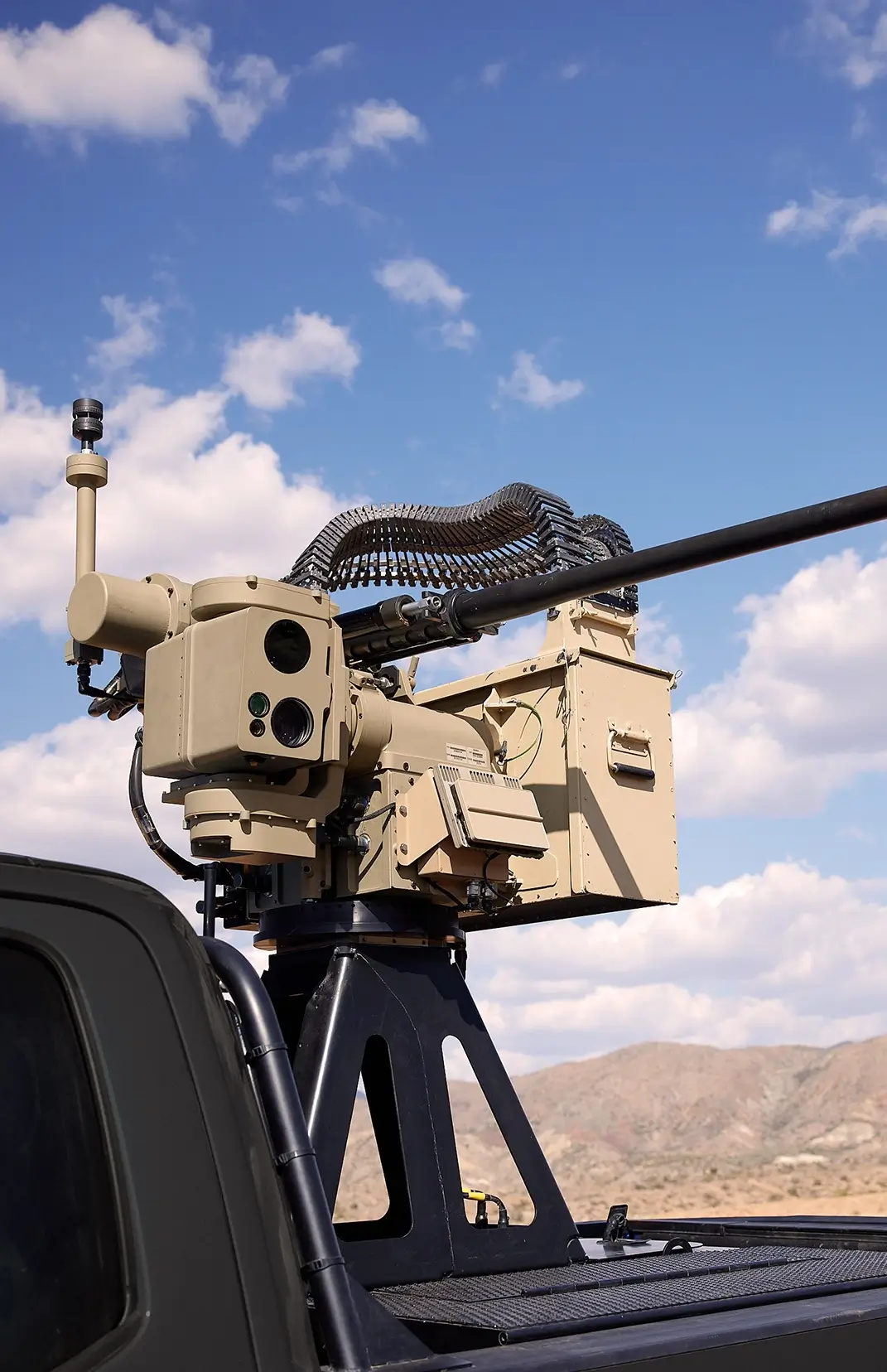 Mounted military remote weapon system with large gun barrel and linked ammunition belt on vehicle under blue sky.