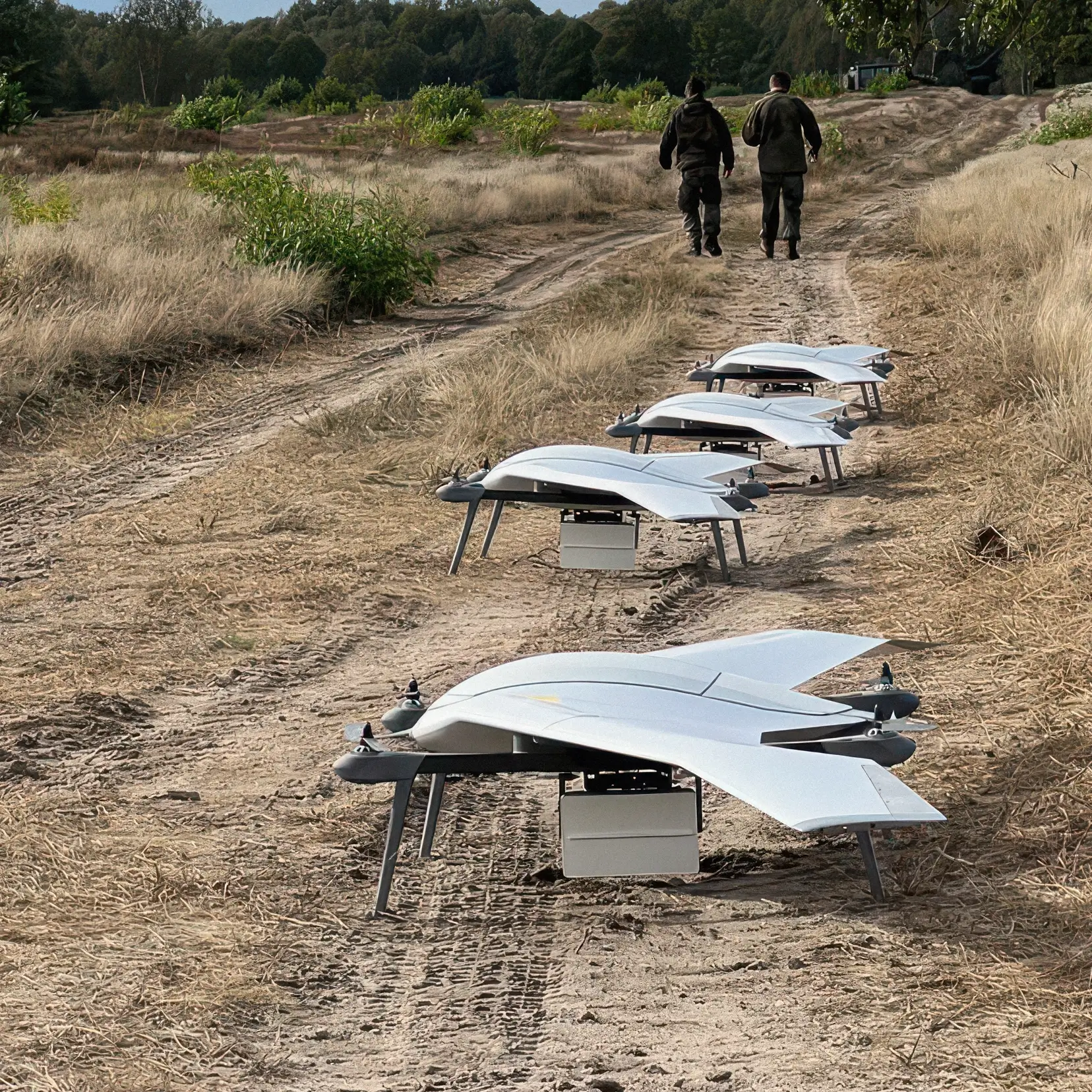 Four white drone aircraft with airborne radar lined up on a dirt path in a dry grassy field.