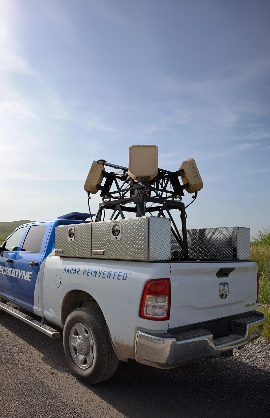 White and blue pickup truck with radar equipment and storage boxes on the back under a clear sky.