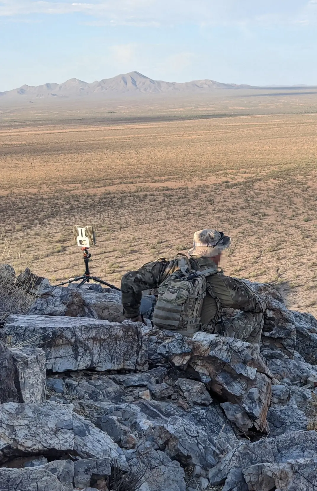 Soldier in camouflage sitting on rocky terrain overlooking vast desert landscape and distant mountains.