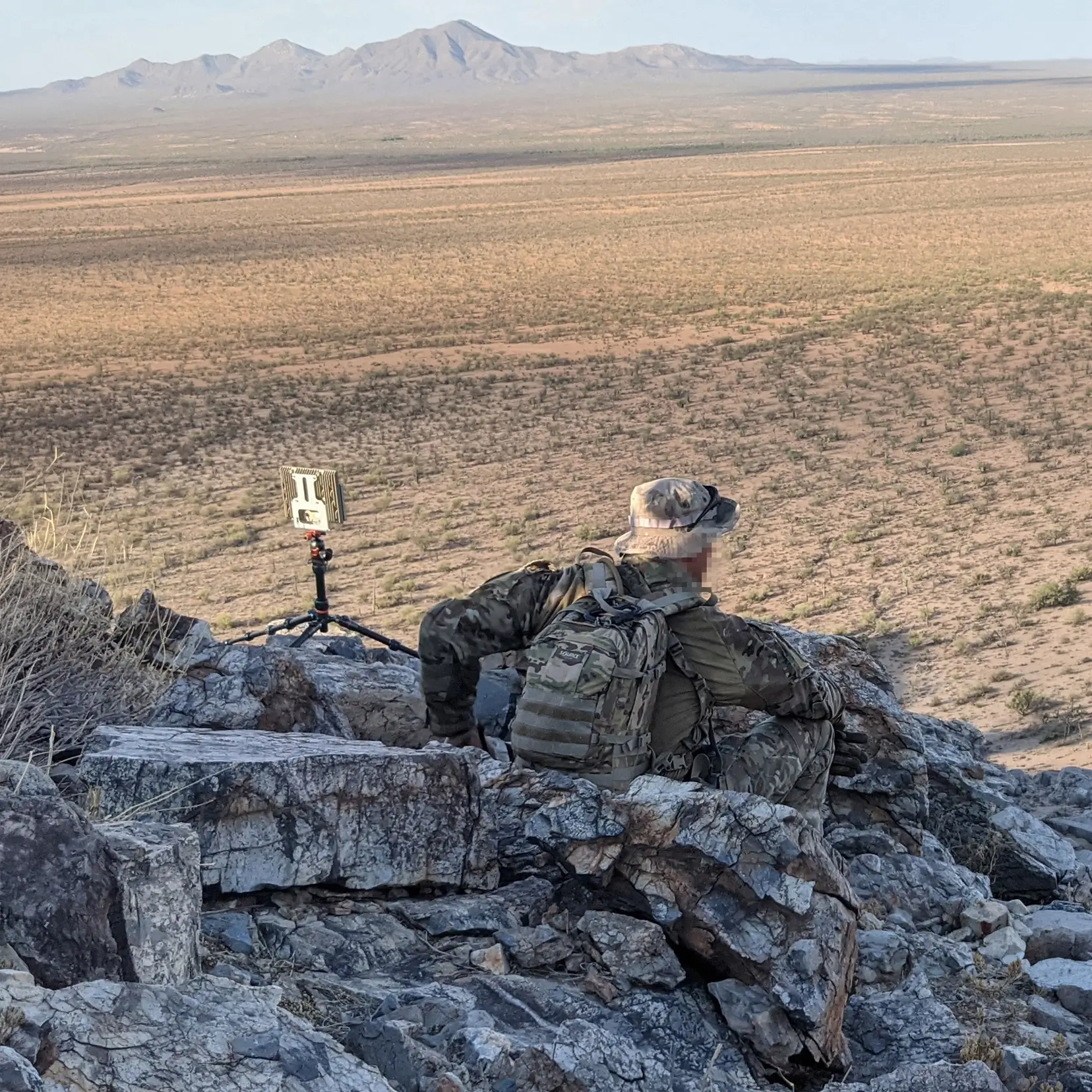 Soldier in camouflage sitting on rocky terrain overlooking a vast desert landscape and mountains.
