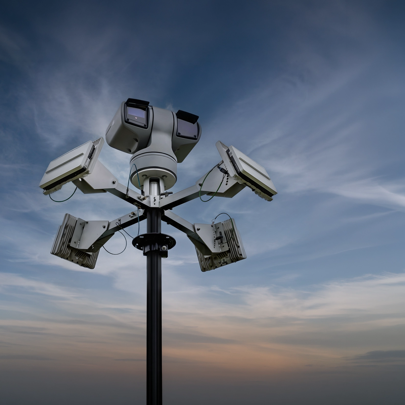 Surveillance camera pole with multiple cameras and lights under a cloudy sky at dusk.