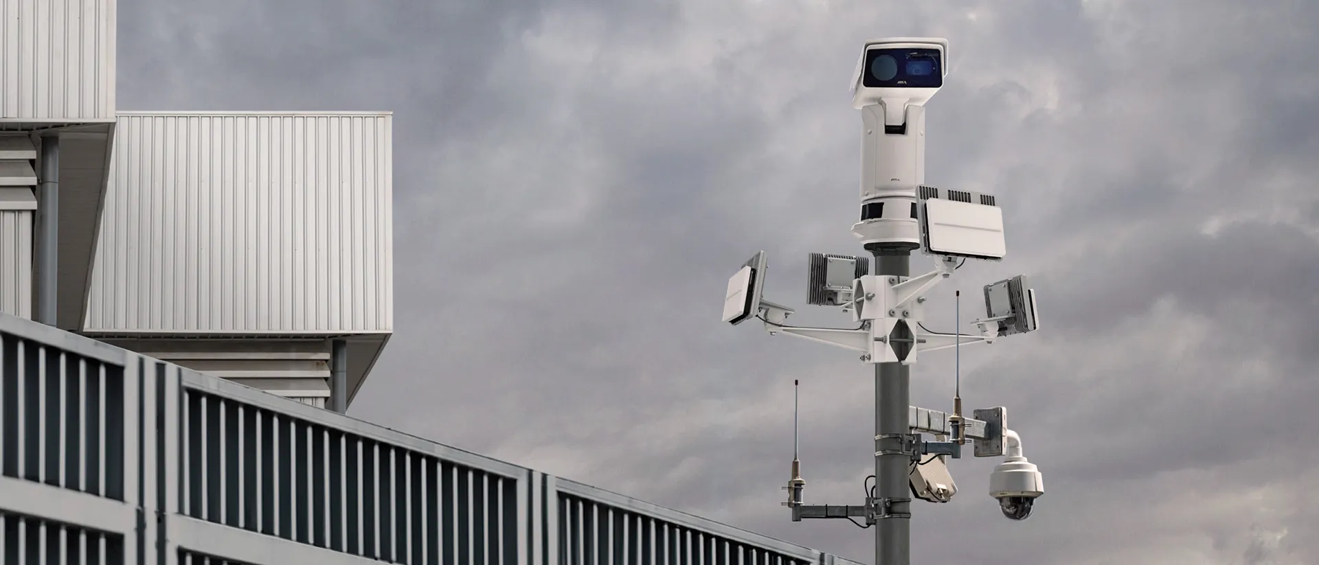 Surveillance tower with multiple cameras and antennas against a cloudy sky near a building.
