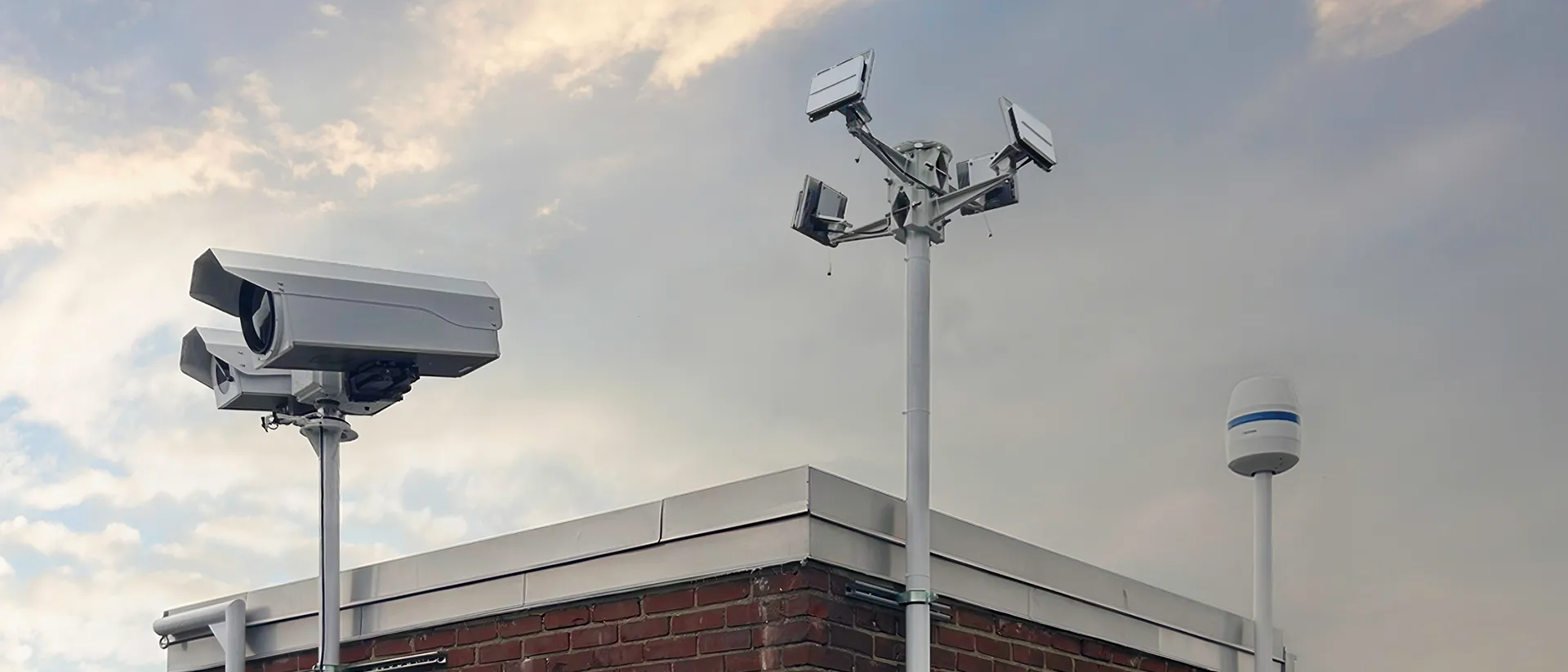 Three different surveillance cameras mounted on poles atop a brick building under a cloudy sky.