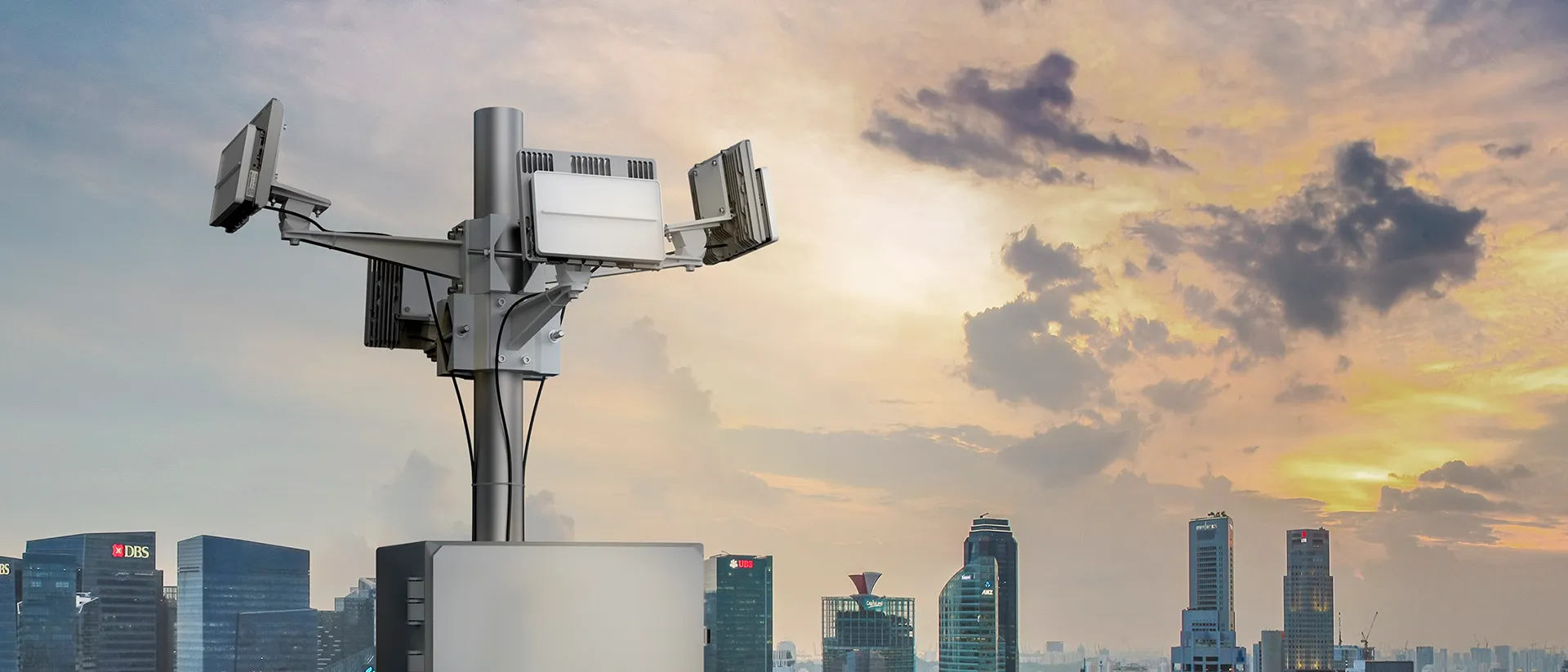 Gray telecom tower with multiple antennas against a city skyline and sunset sky.