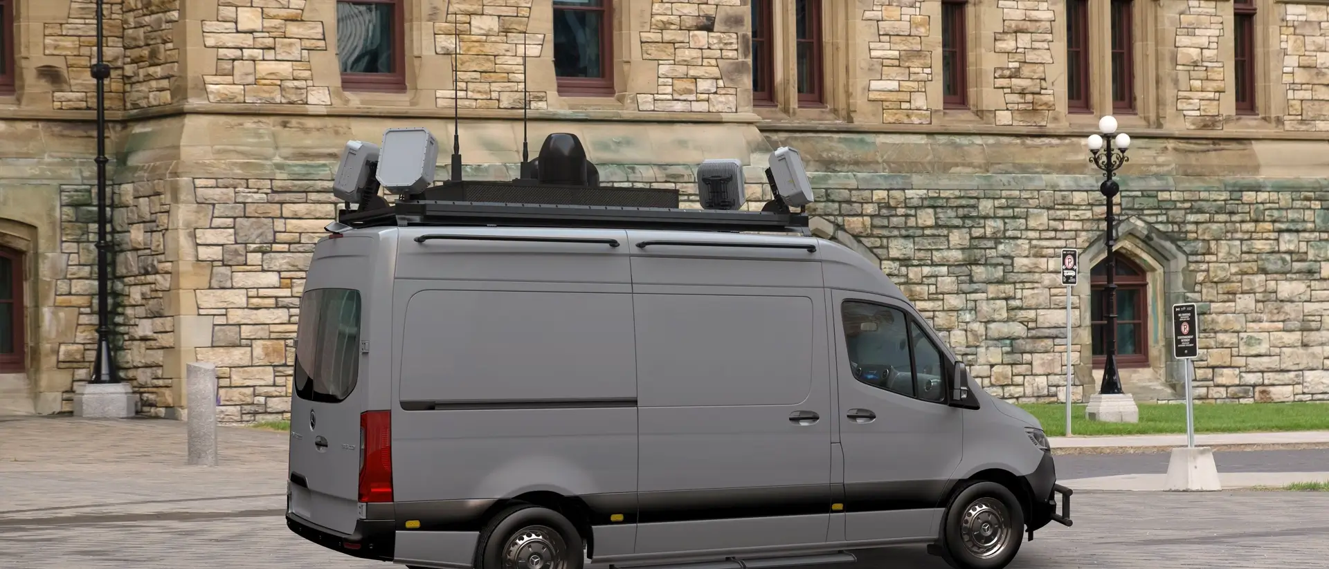 Gray van equipped with roof-mounted radar and communication devices parked near a stone building.