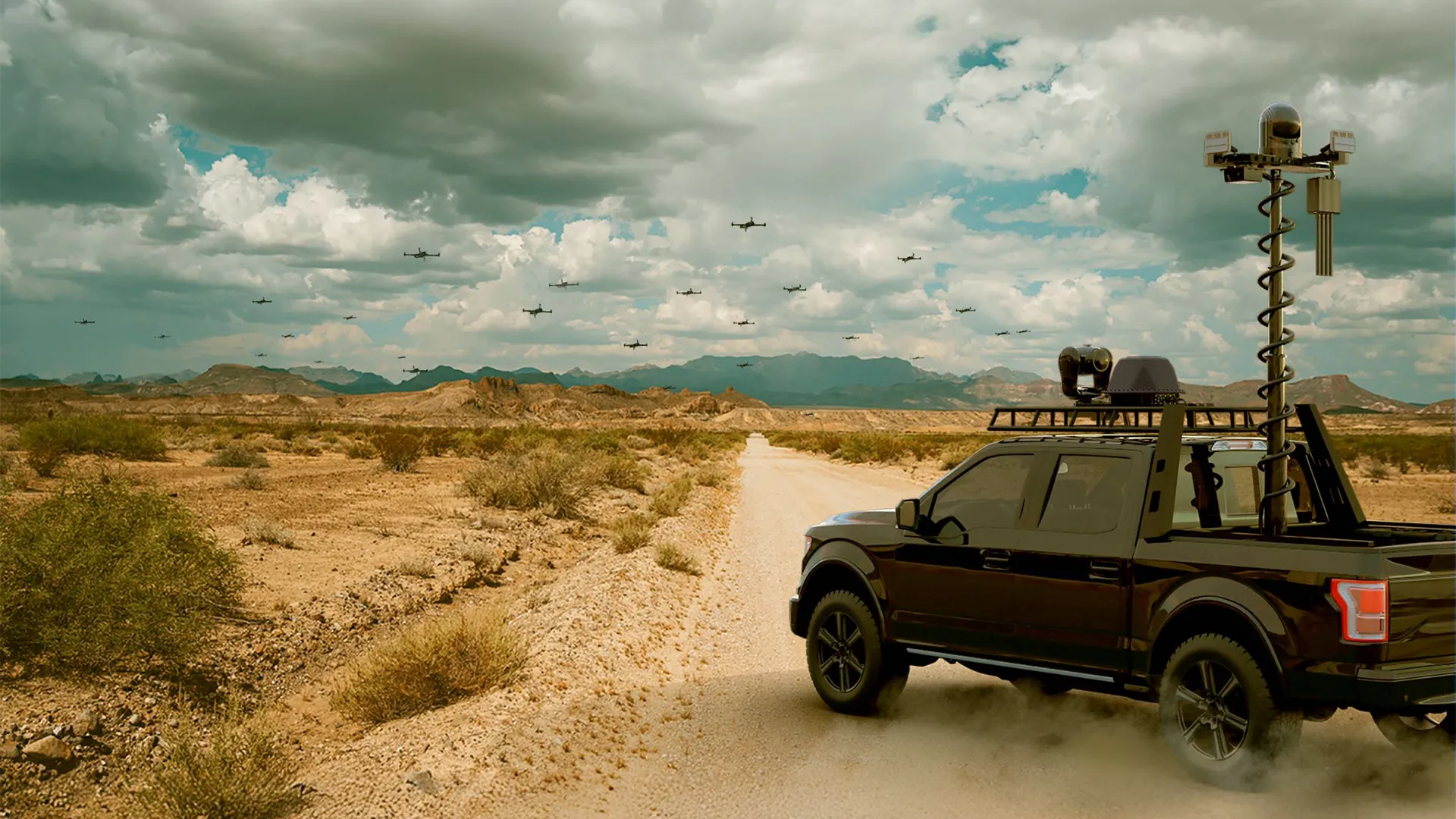 Black pickup truck with mounted sensors drives on desert road under cloudy sky with flying drones.