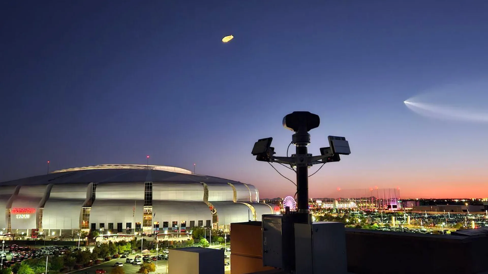 State Farm Stadium at dusk with a security camera tower in the foreground and a Ferris wheel in the distance.