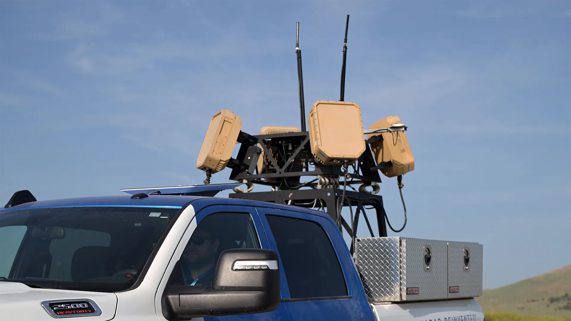 Blue and white heavy duty truck with radar and antenna equipment mounted on its roof.