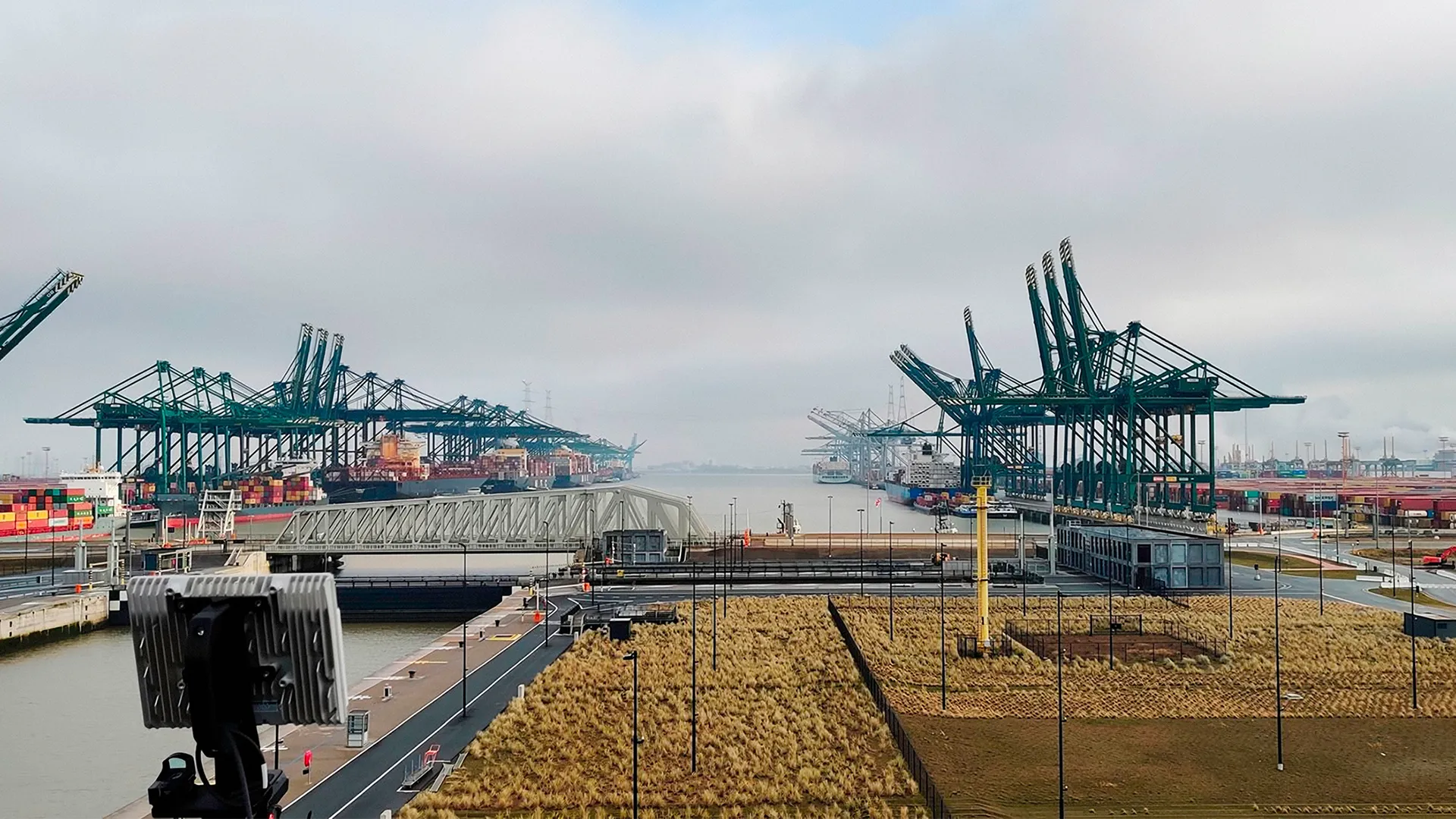View of a busy shipping port with cranes, container ships, and stacked cargo containers under a cloudy sky.