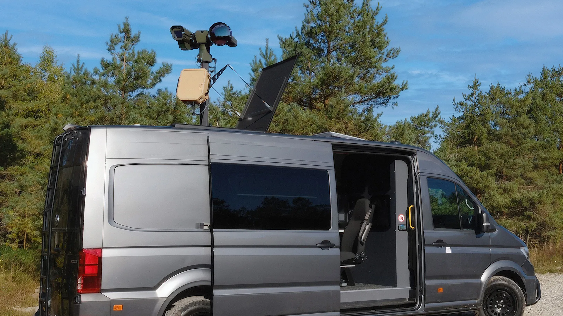 Gray surveillance van with open side door and equipment mounted on roof in outdoor setting.