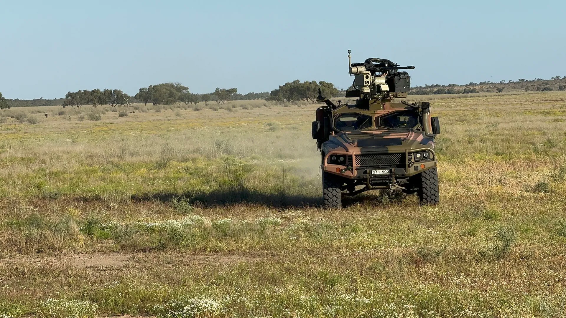 Military armored vehicle with mounted weapon driving on grassy plain under blue sky.