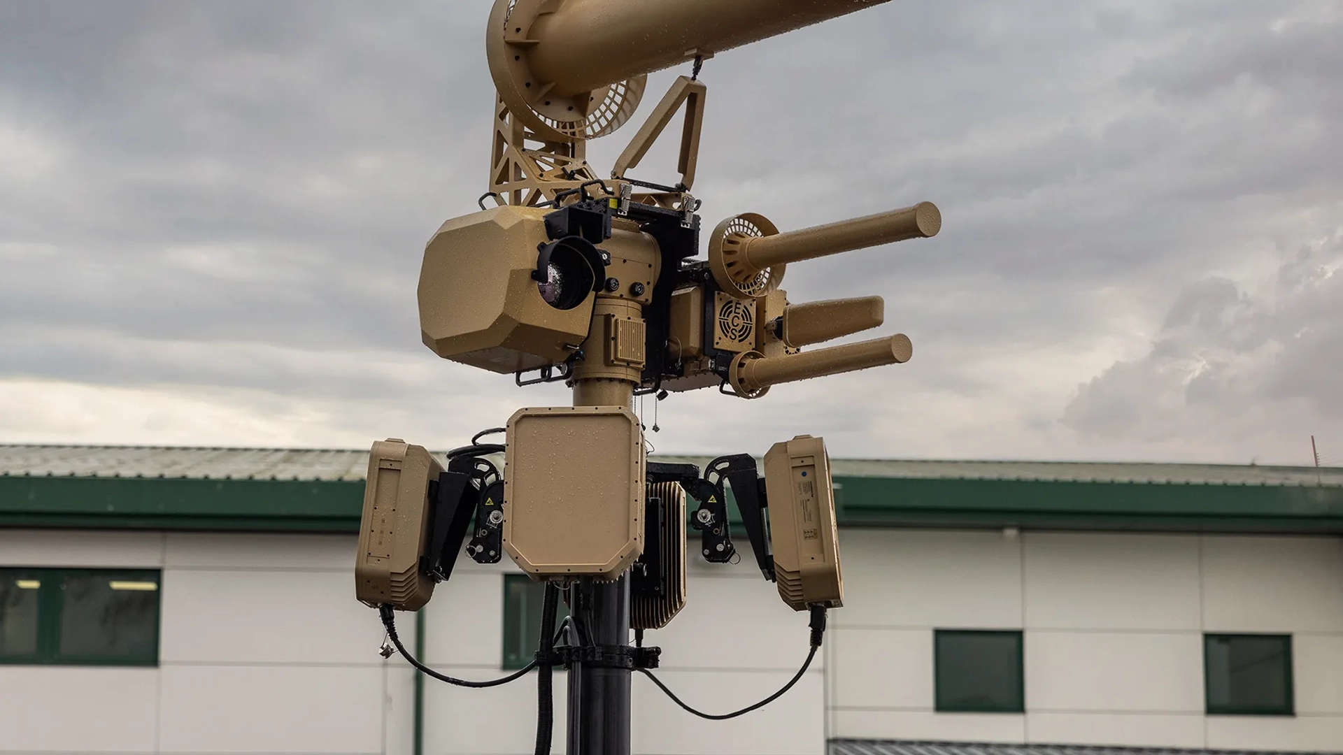 Mounted tan-colored multi-barrel security or defense system with cameras on a pole outside a building.