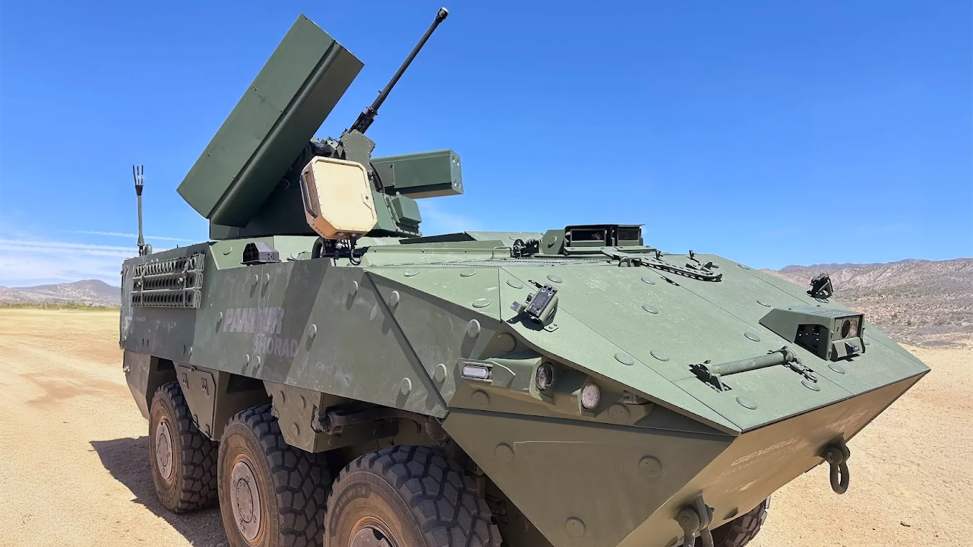Green armored military vehicle with six large wheels and mounted weapons under a clear blue sky.
