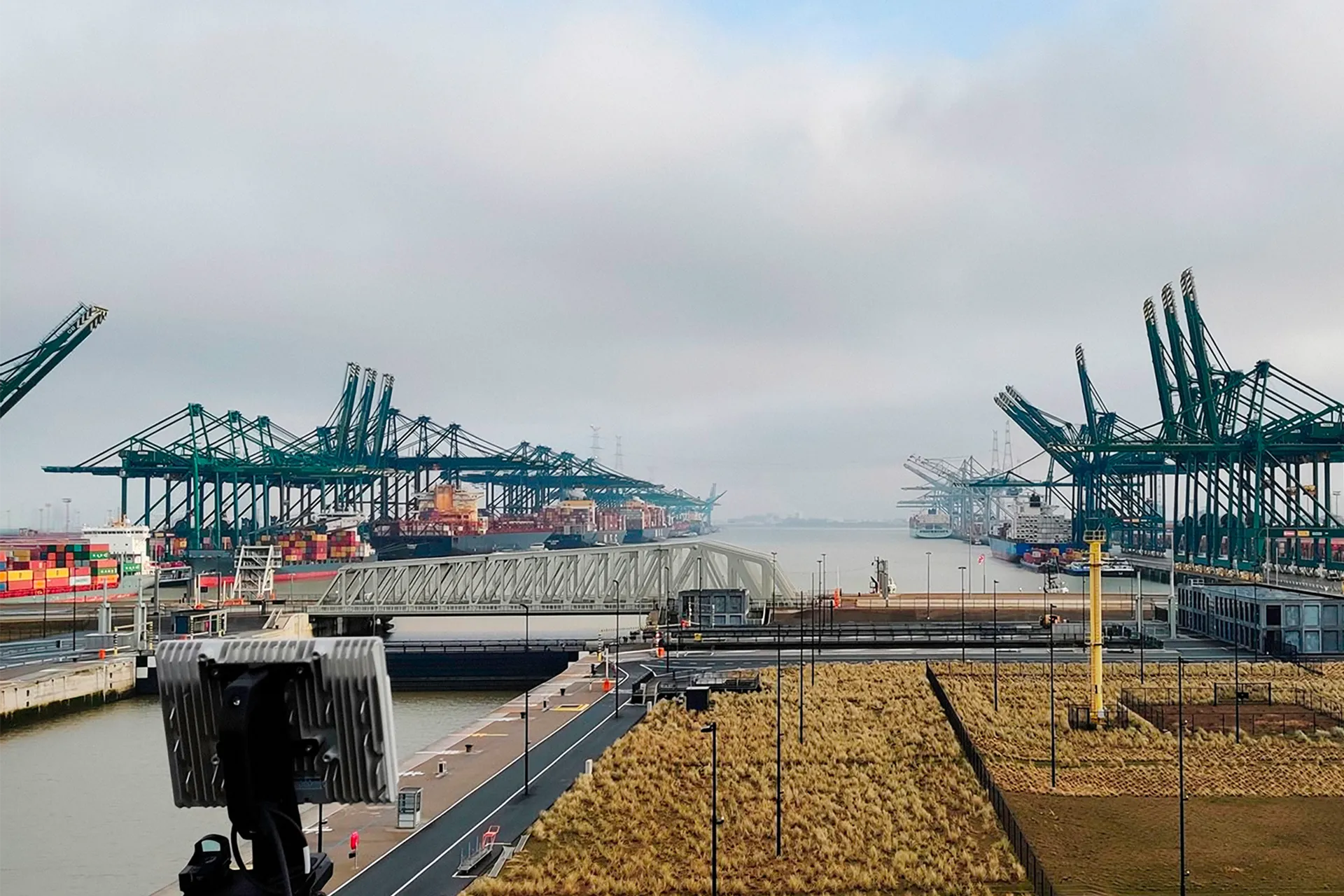 View of a busy shipping port with cranes, container ships, and a waterway under a cloudy sky.