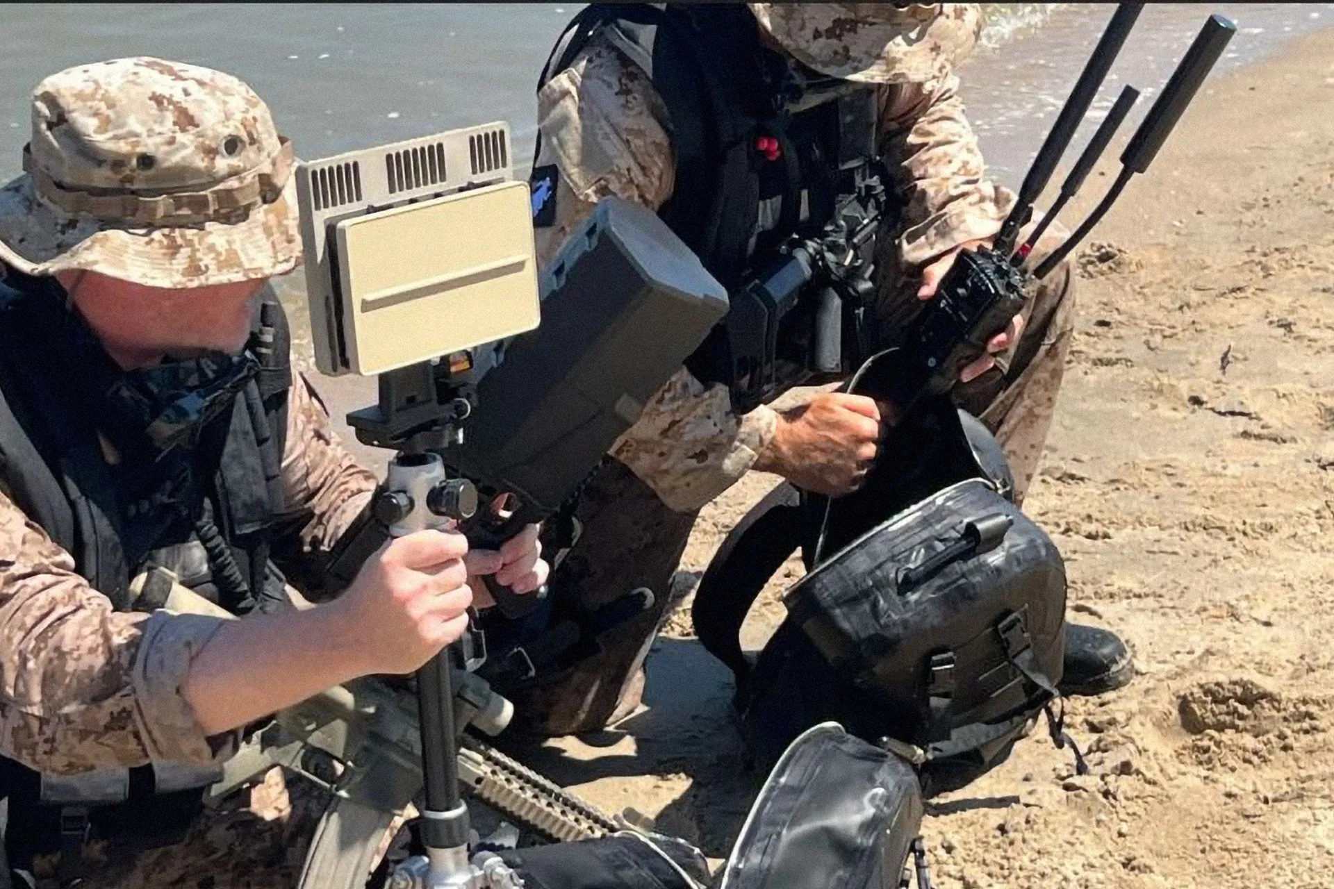 Two soldiers in camouflage kneeling on sand, handling electronic equipment with antennas.