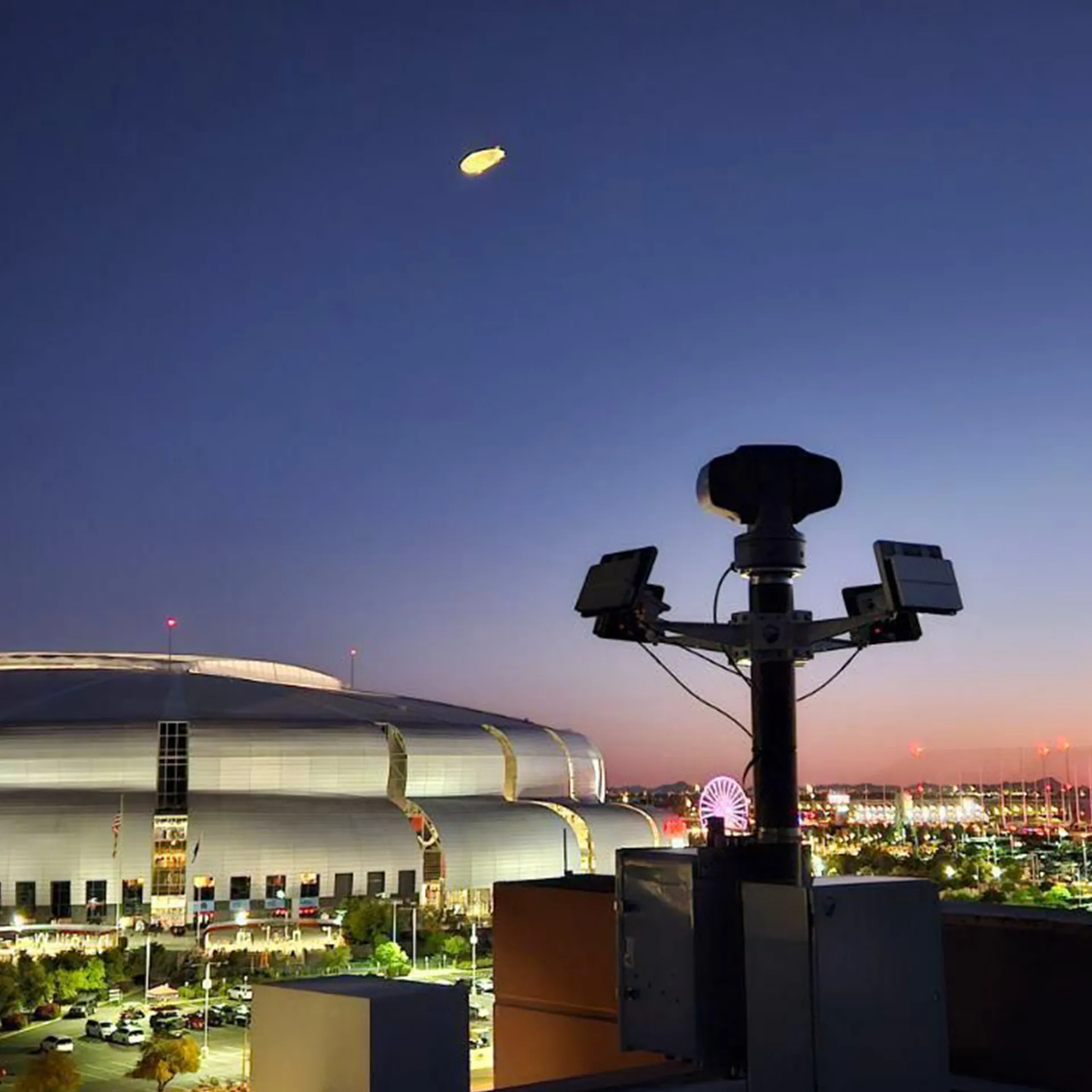 Security camera system overlooking a lit stadium at dusk with a blimp and city lights in background.