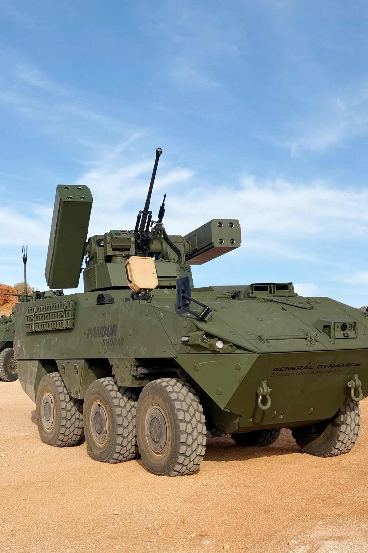 Green Pandur SHORAD armored vehicle with missile launchers and mounted gun on sandy terrain under blue sky.