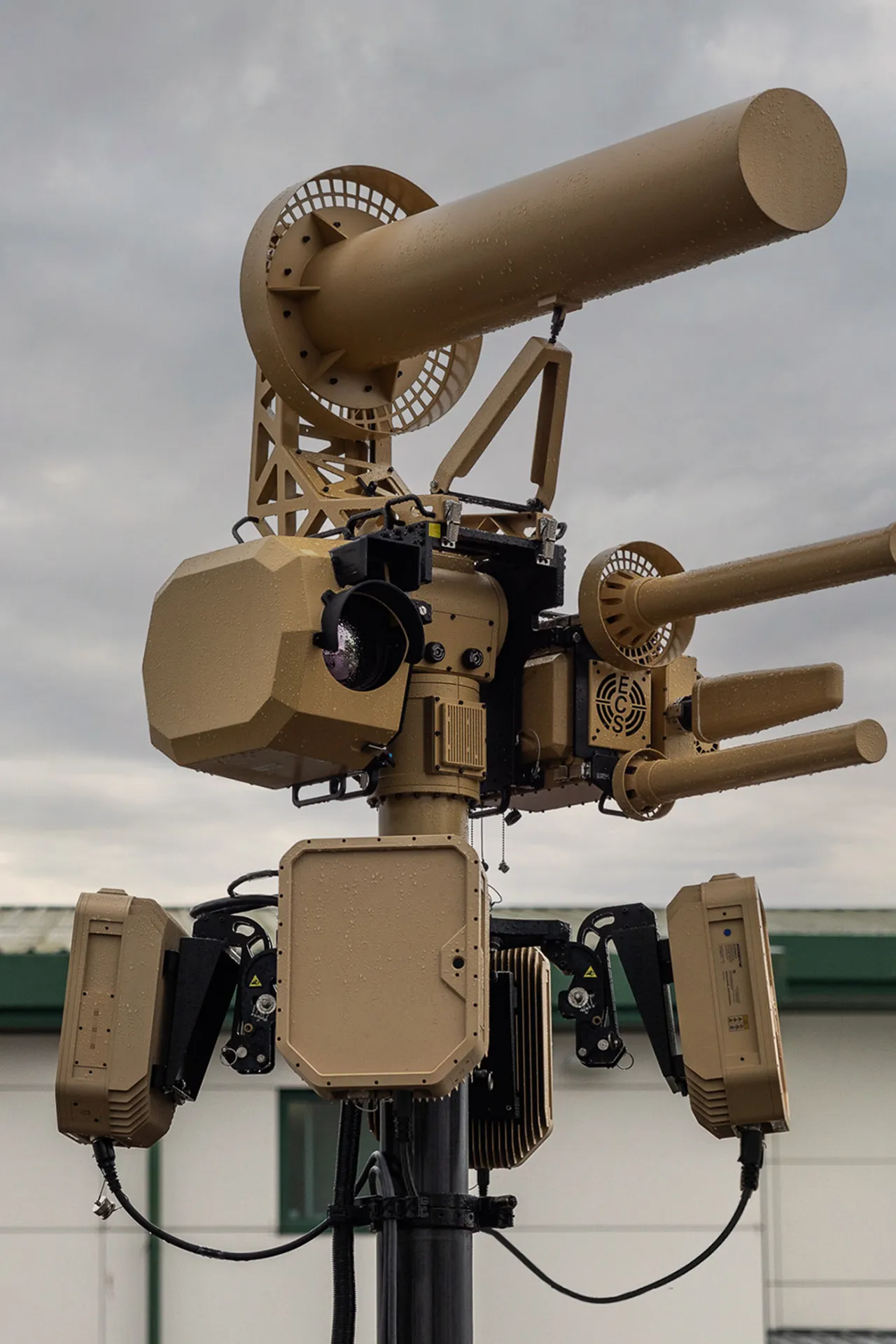 Close-up of a tan robotic defense system with multiple barrels mounted on a black pole outdoors.