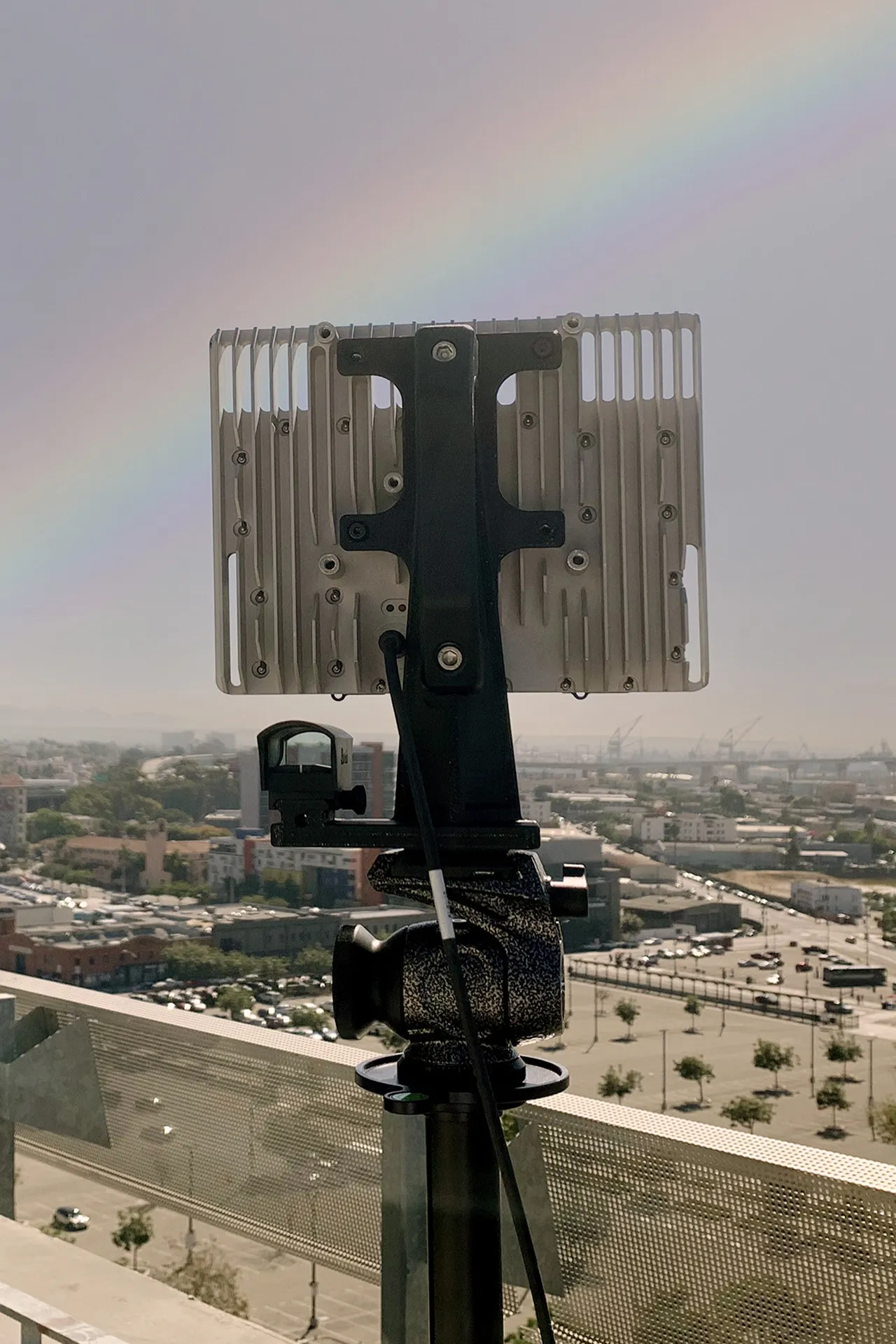 Rear view of a mounted electronic device with cooling fins overlooking a cityscape and a faint rainbow.