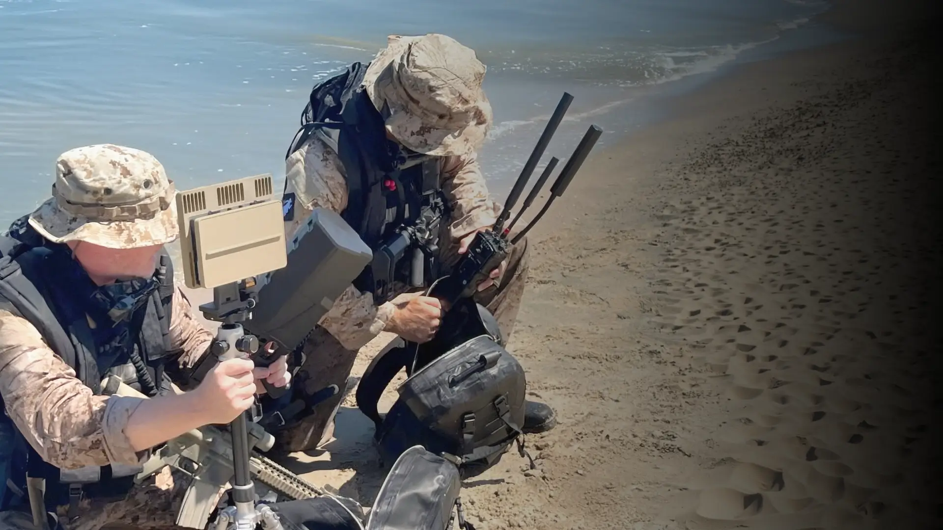 Two soldiers in camouflage gear kneeling on a sandy beach with communication equipment.