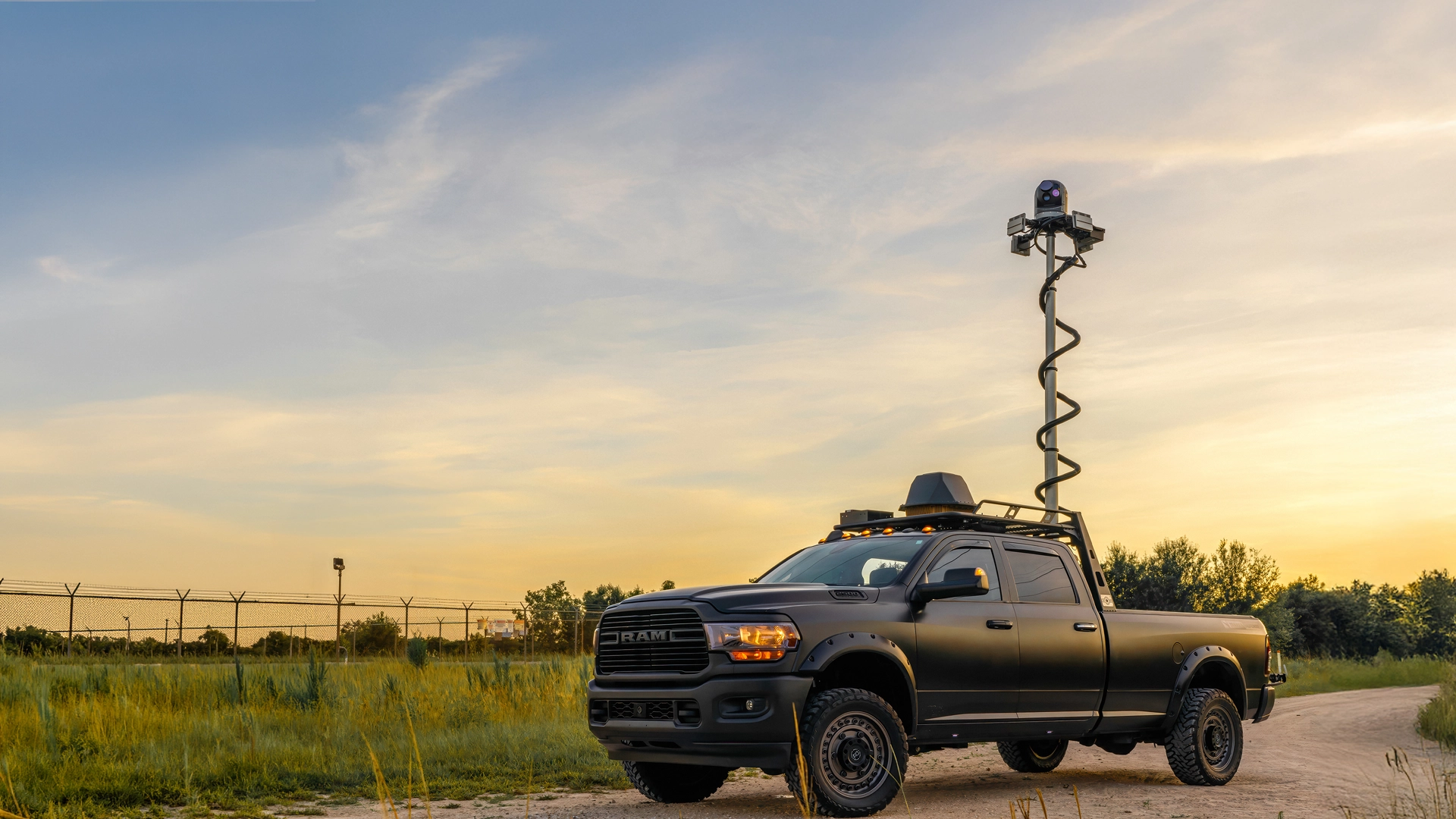 Black RAM pickup truck with raised camera mast parked on dirt road at sunset.