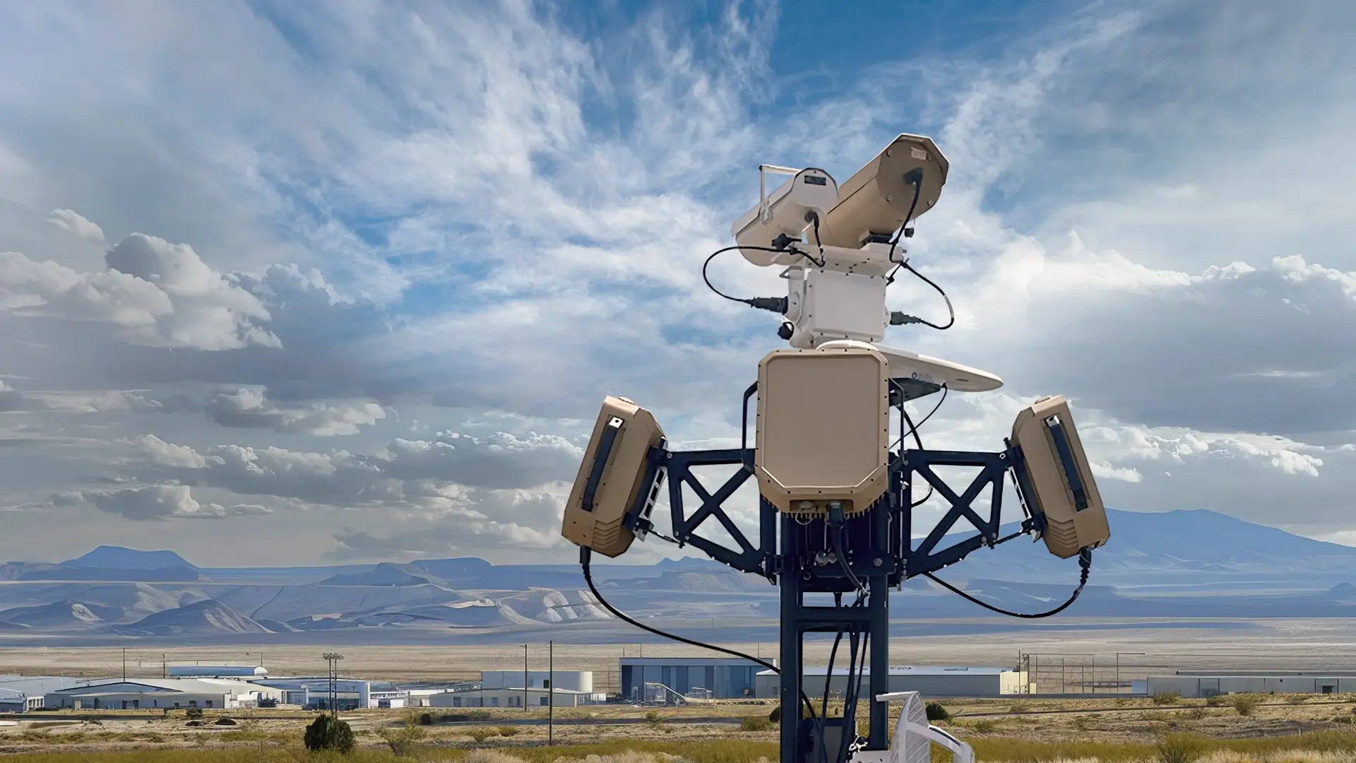 Security sensor tower with various mounted devices in a desert area with buildings and mountains behind.