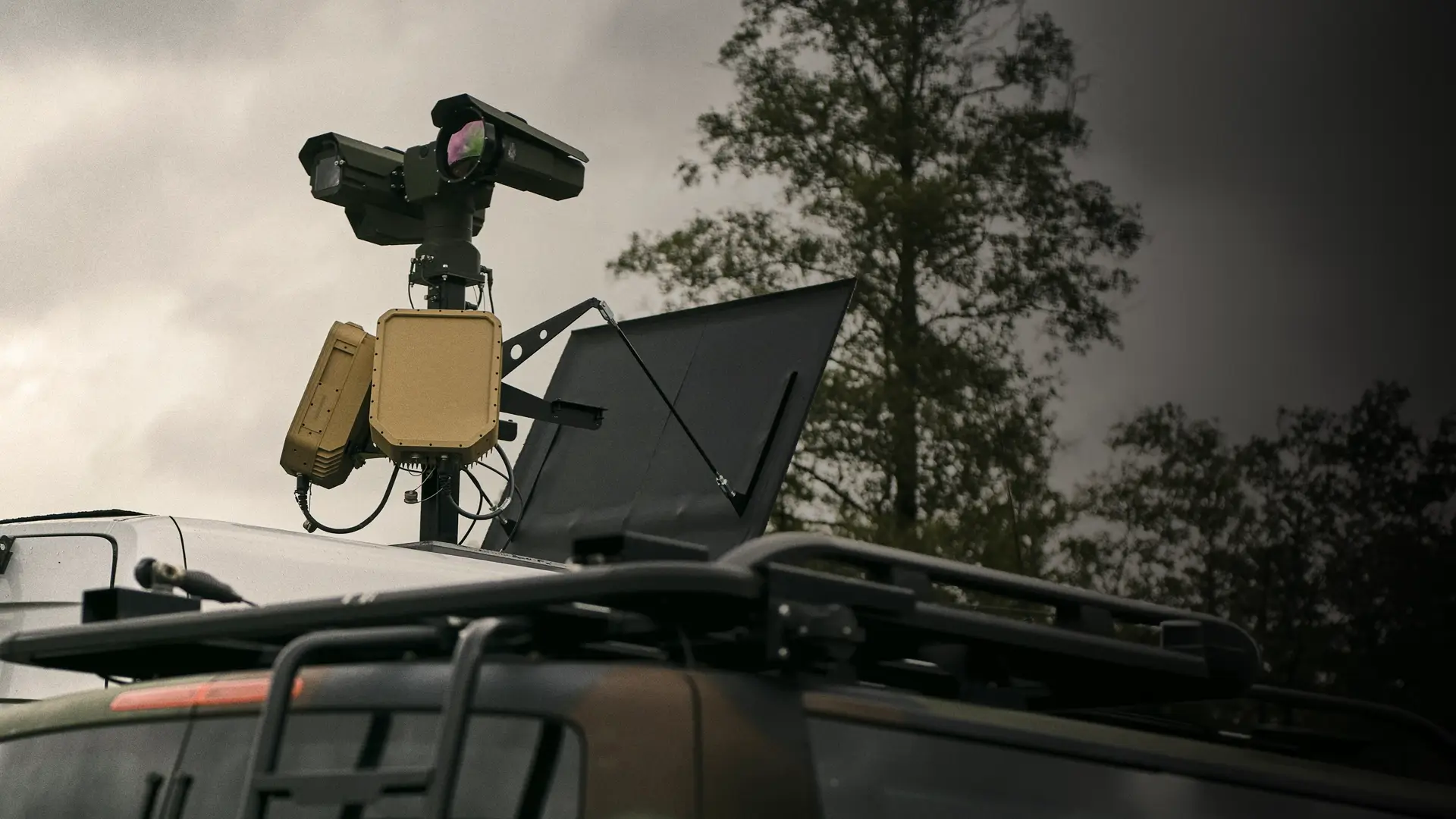 Surveillance equipment mounted on top of a vehicle against a cloudy sky and trees.