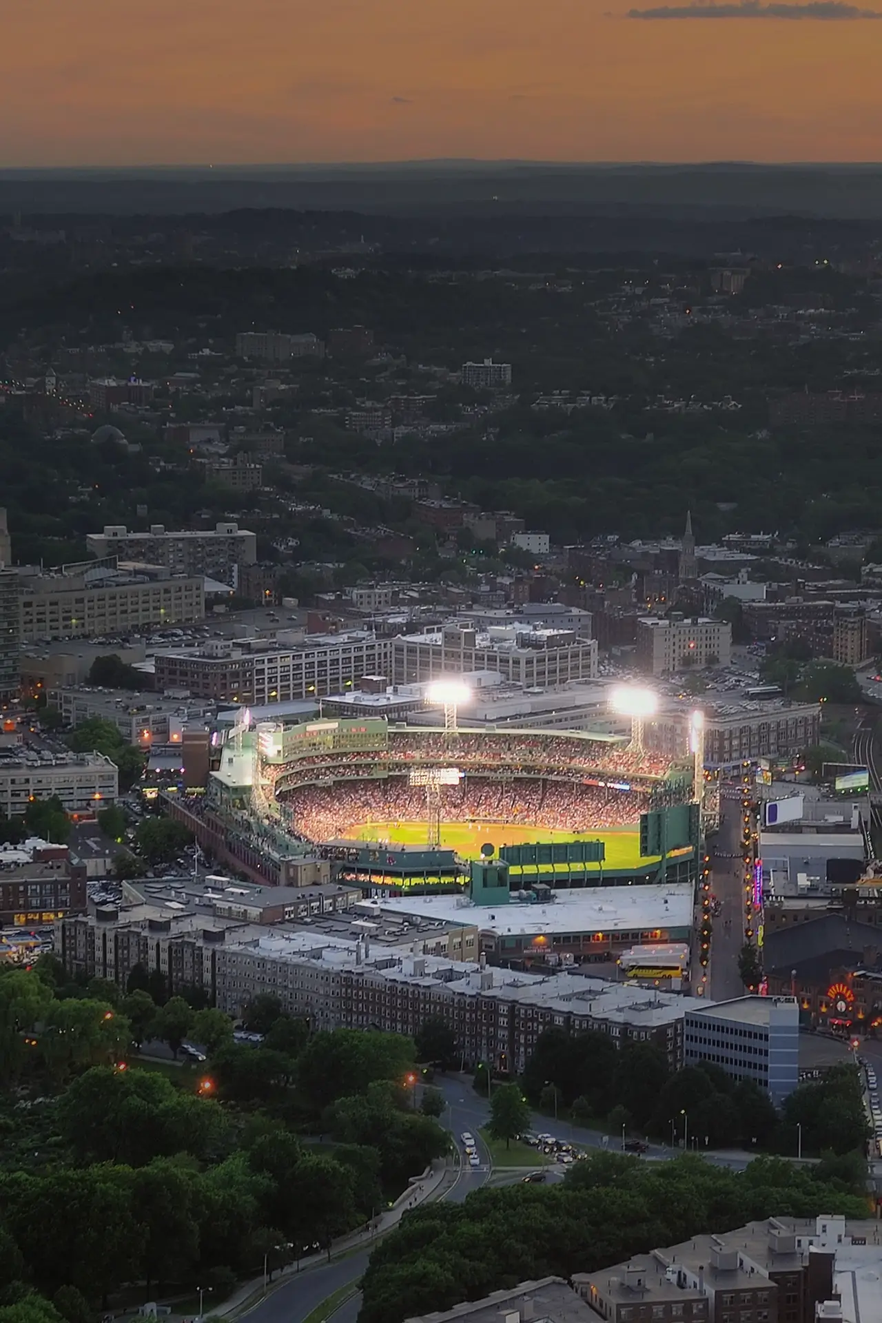 Aerial view of a brightly lit baseball stadium at dusk within a city surrounded by buildings and trees.