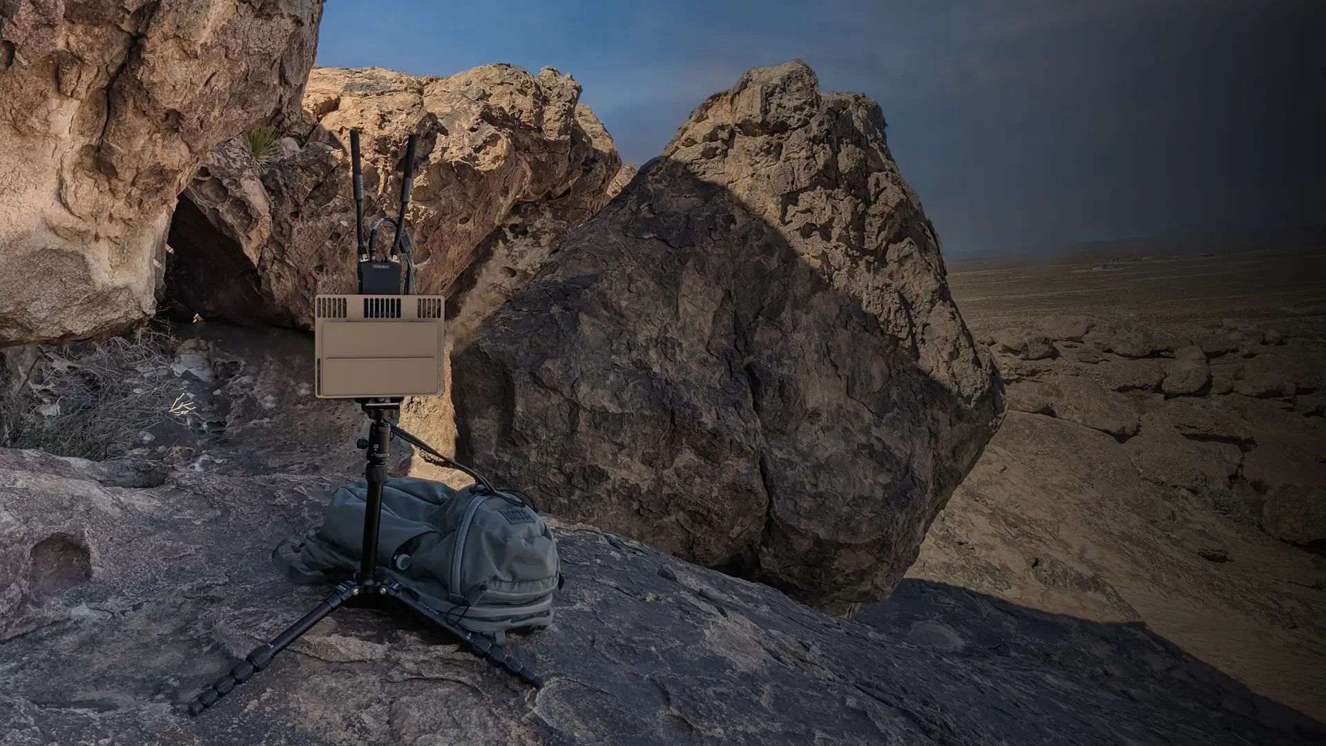 Electronic device with antennas on tripod and backpack among large rocks in a desert-like area.