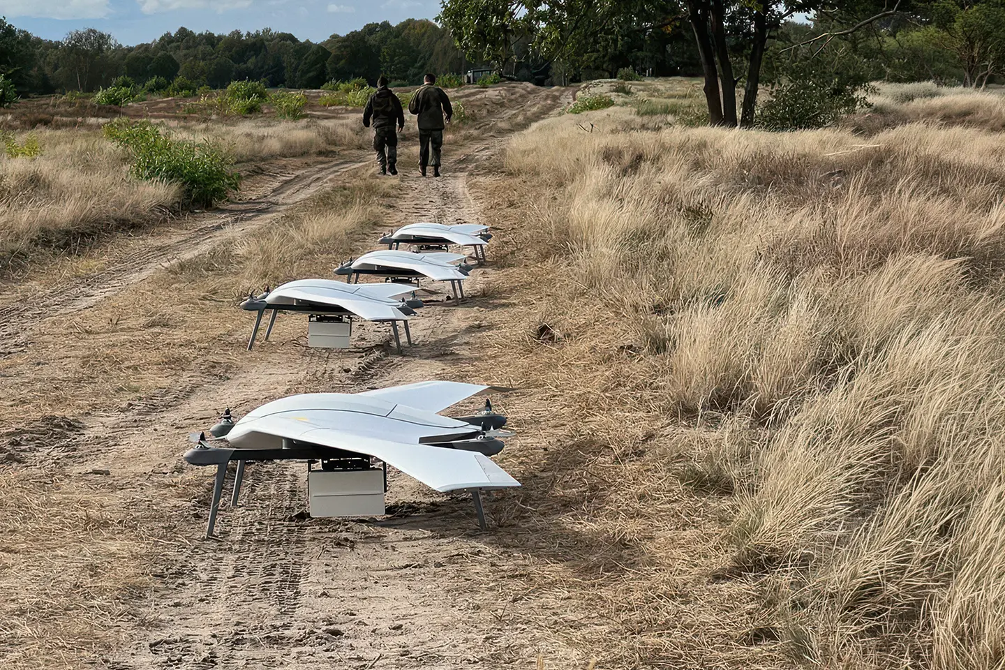Four white drone aircraft with wings and legs lined up on a dirt path in a grassy field.