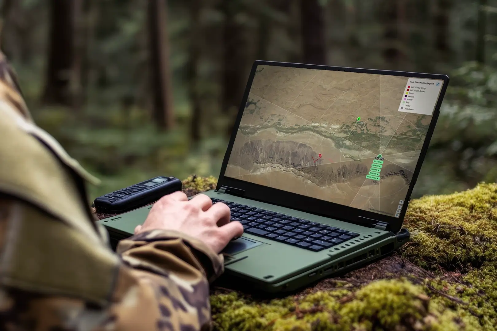 Person in camouflage using rugged laptop outdoors showing a map with tracking markers on screen.