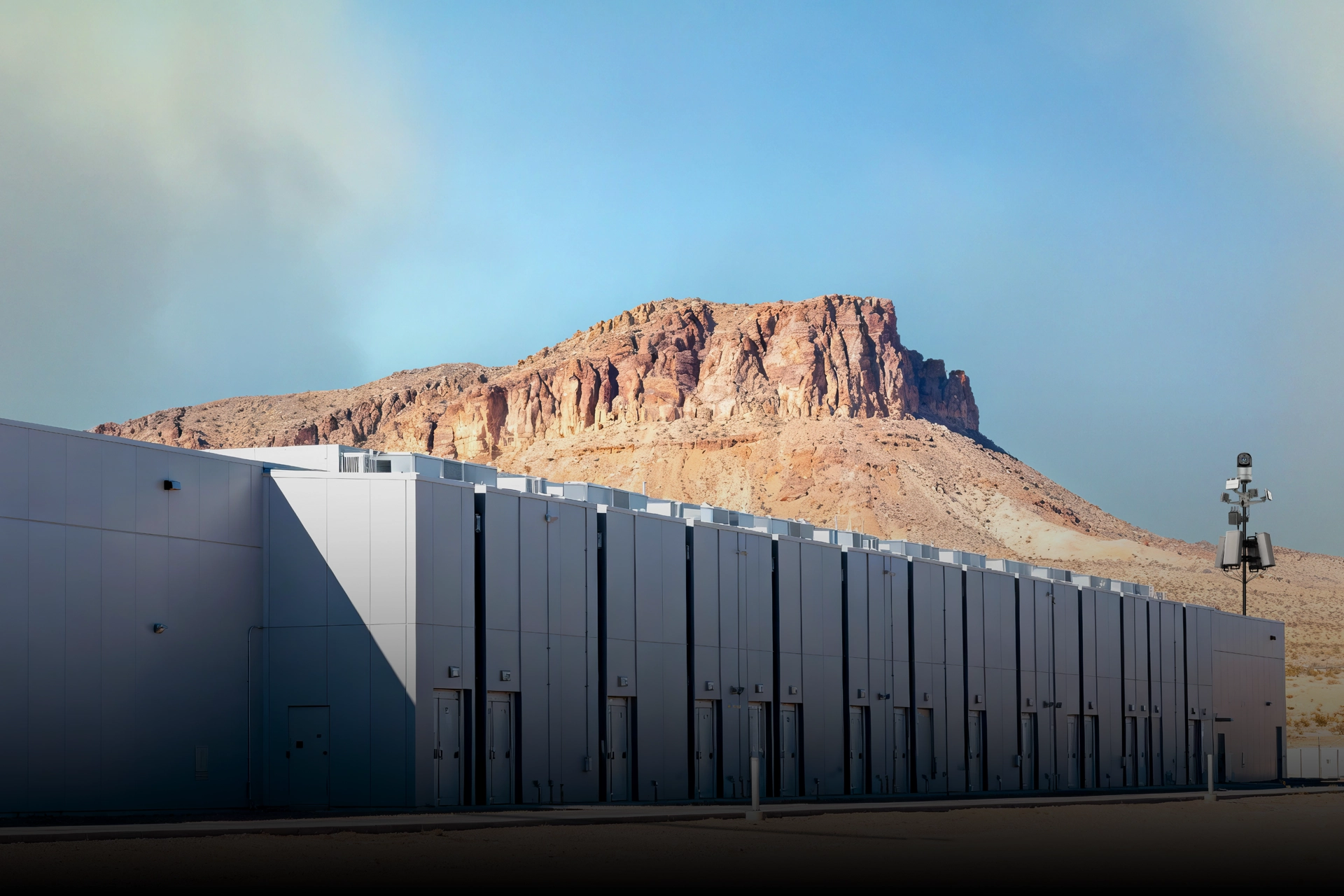 Large modern data center building with security cameras and a rocky desert mountain in the background.