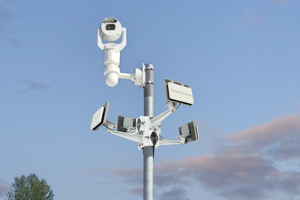 Security camera and floodlights mounted on a tall metal pole against a blue sky with clouds.