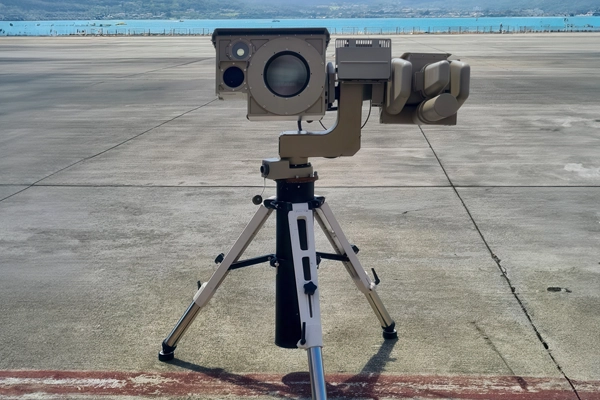 Tripod-mounted surveillance camera set up on a concrete surface near a body of water.