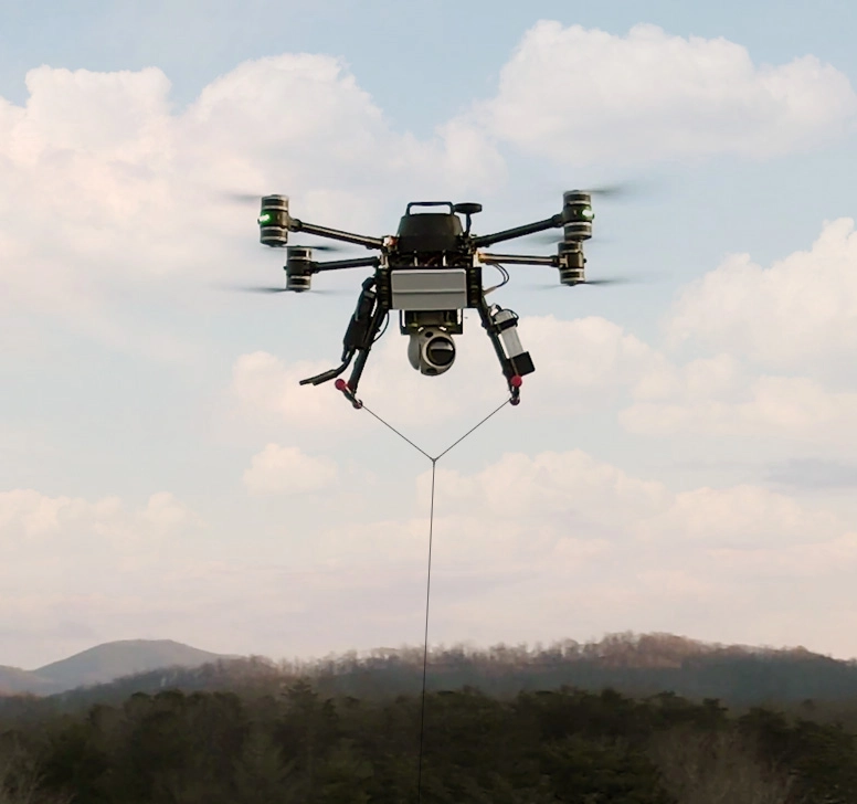 Black drone with camera attached, hovering in cloudy sky above forested hills.
