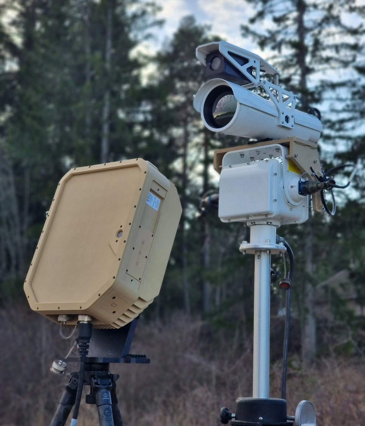 Two security devices on tripods outdoors with trees in the blurred background.