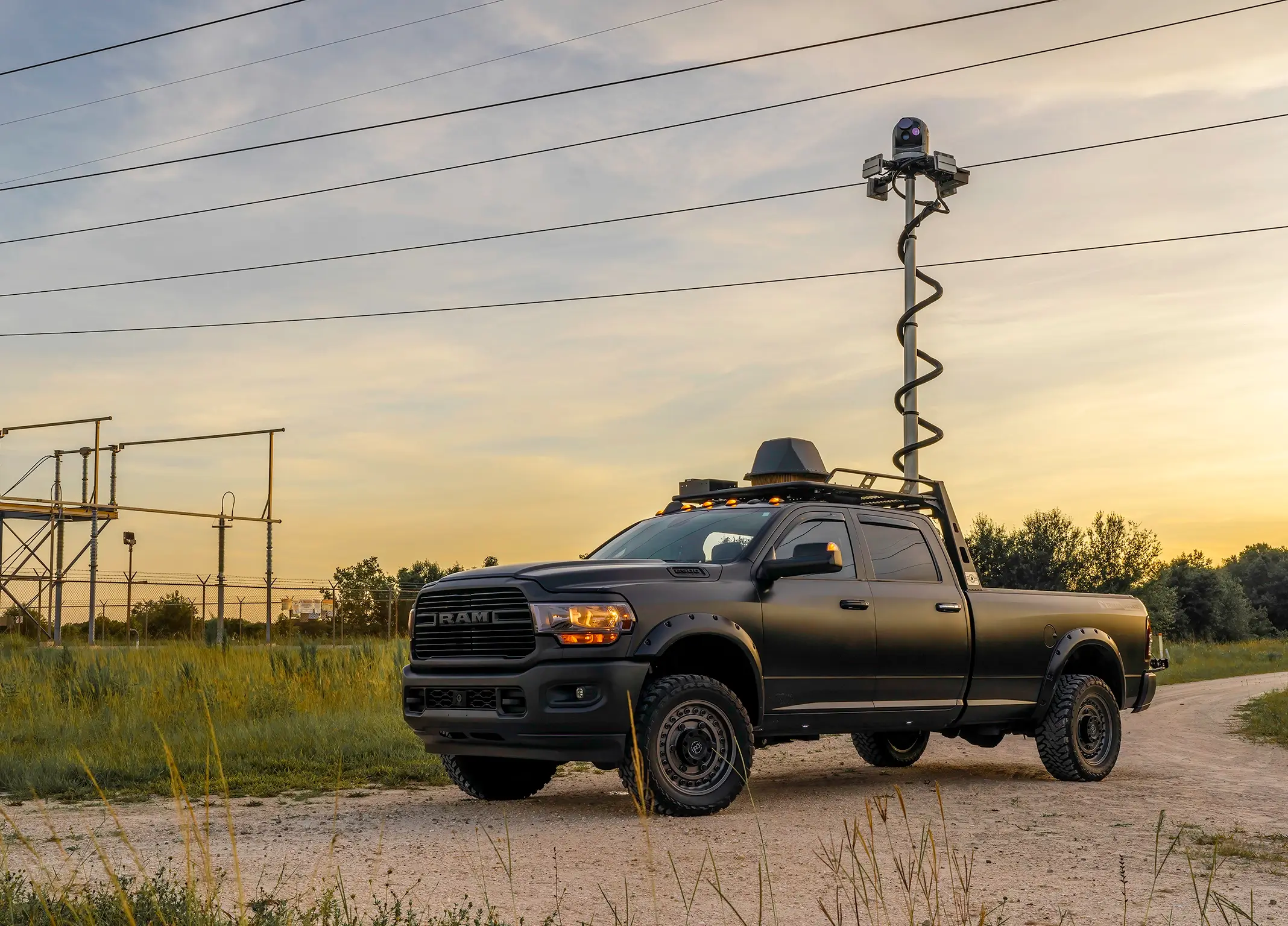 Black RAM pickup truck with raised surveillance camera mast parked on dirt road at sunset.