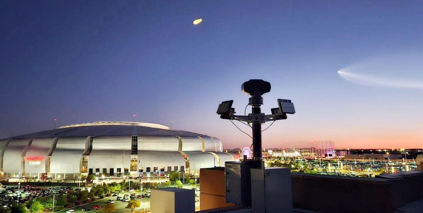 State Farm Stadium at dusk with a camera rig in the foreground and a comet or bright light streak in the sky.