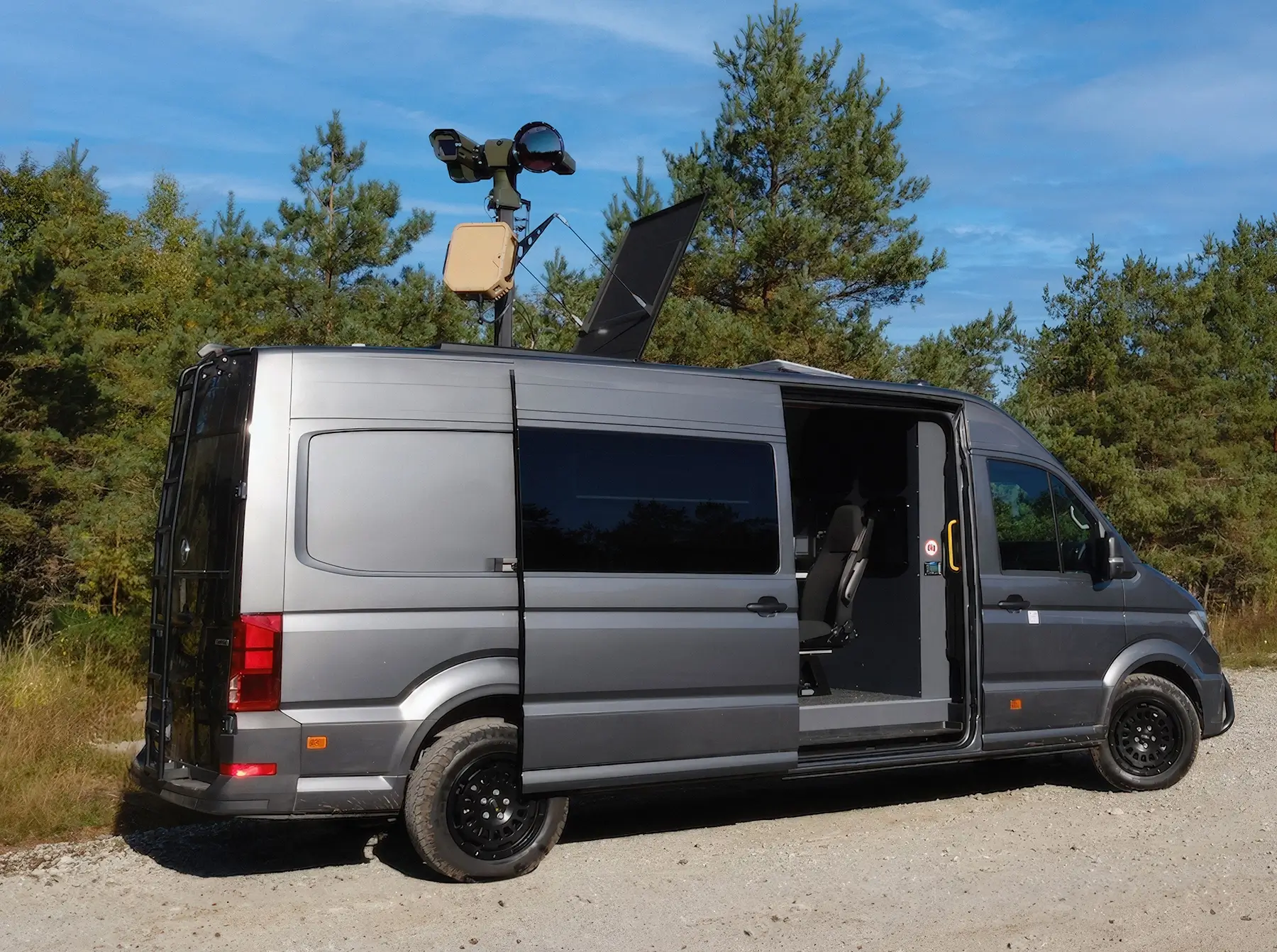 Gray van with open side door and mounted surveillance equipment on roof, parked on gravel near trees.