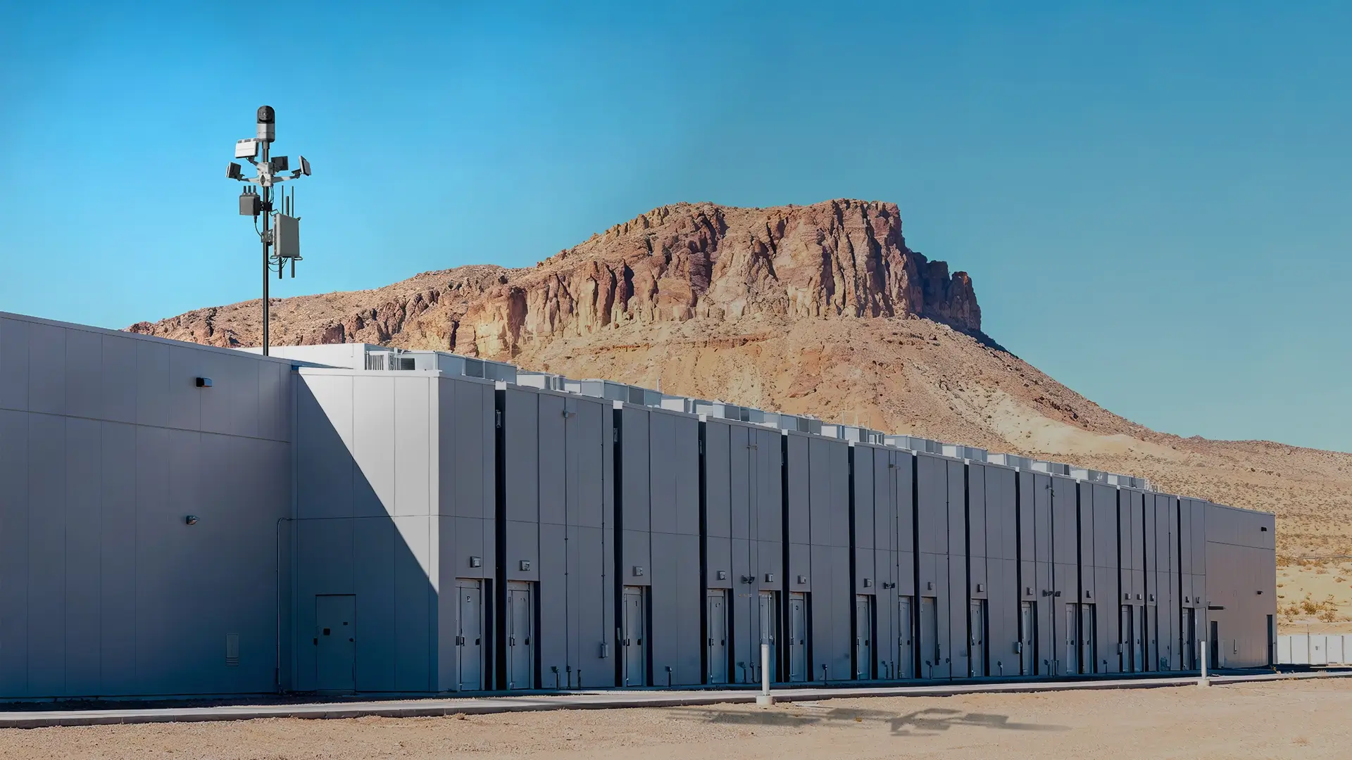 Long gray data center building with multiple doors and security cameras in a desert with a rocky hill.