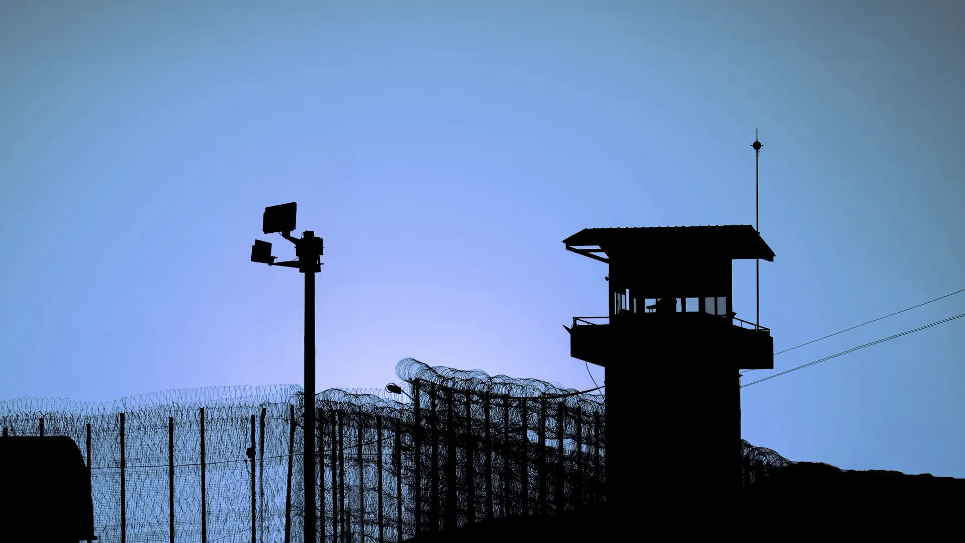 Silhouette of a prison watchtower and barbed wire fence against a blue sky at dusk.