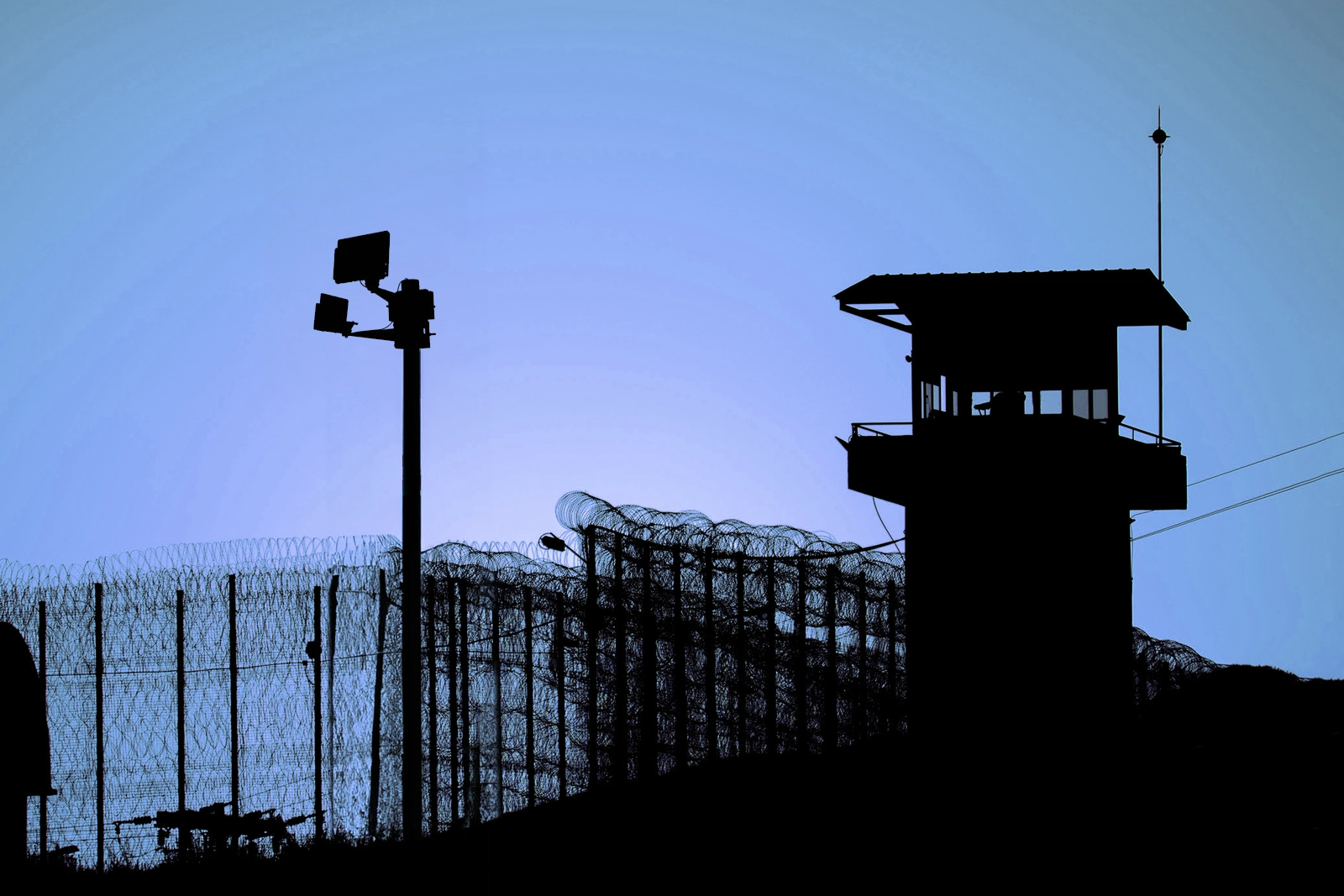 Silhouette of a prison watchtower, barbed wire fence, and security camera under a clear blue sky.
