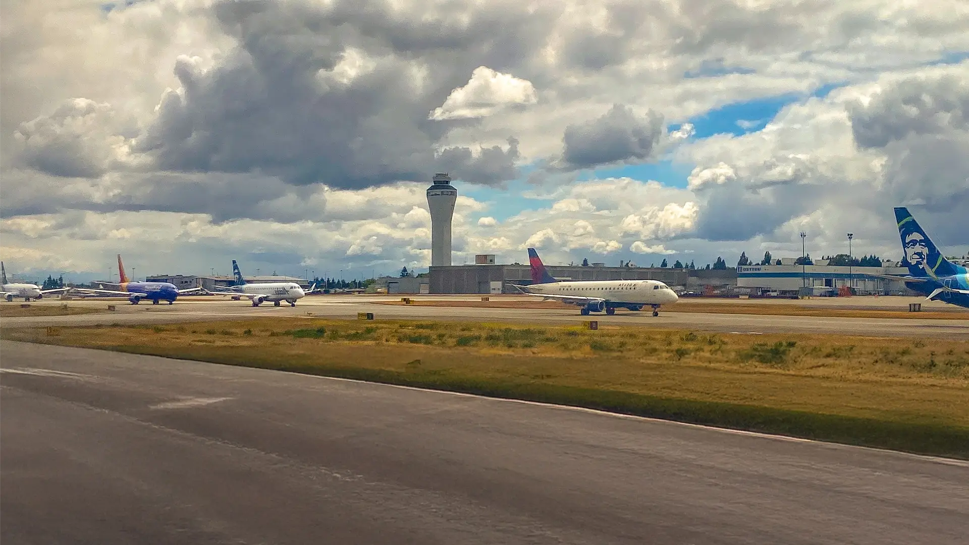 Multiple airplanes are taxiing near an airport control tower under a cloudy sky.