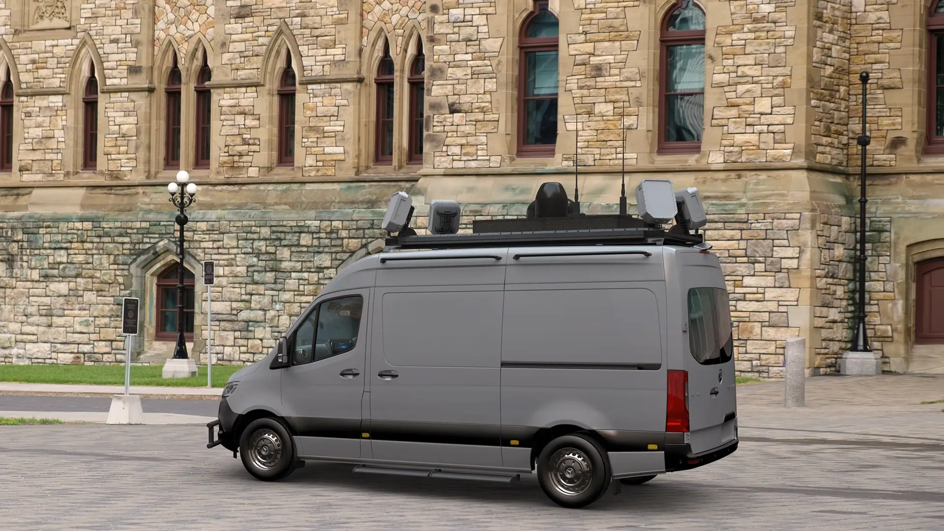 Gray surveillance van equipped with multiple rooftop radar and communication devices parked in front of a stone building.