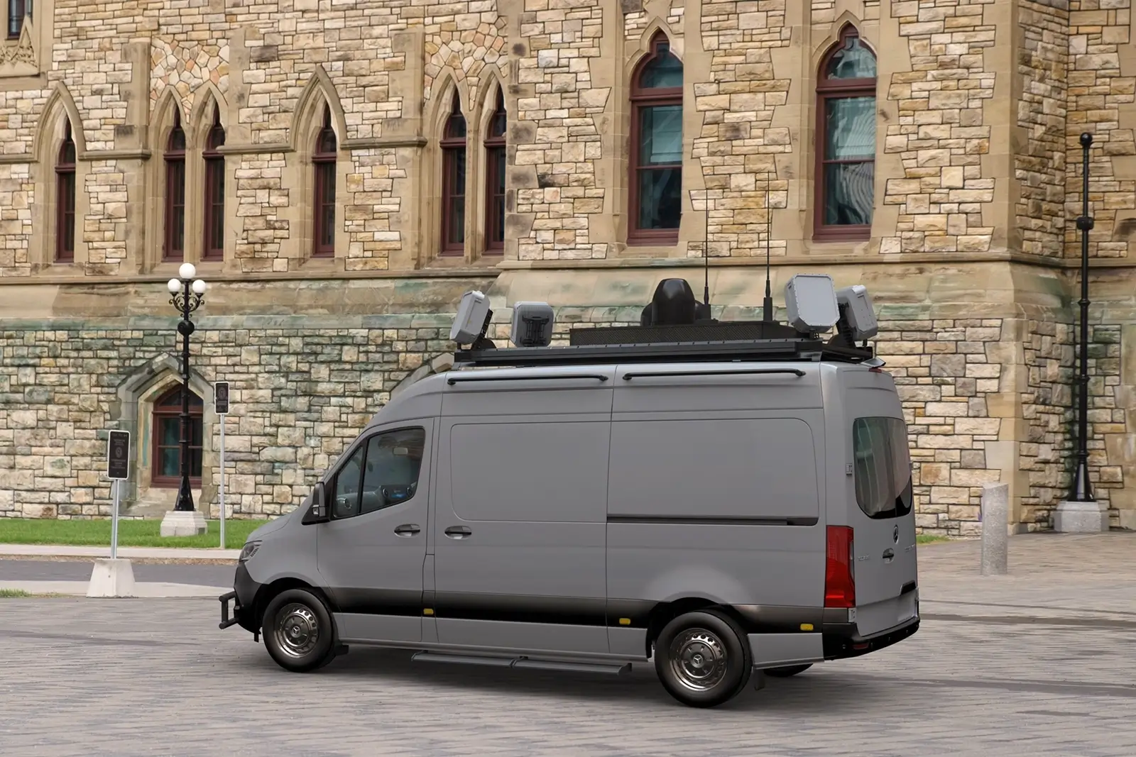 Gray surveillance van with rooftop equipment parked on a paved area near a stone building.