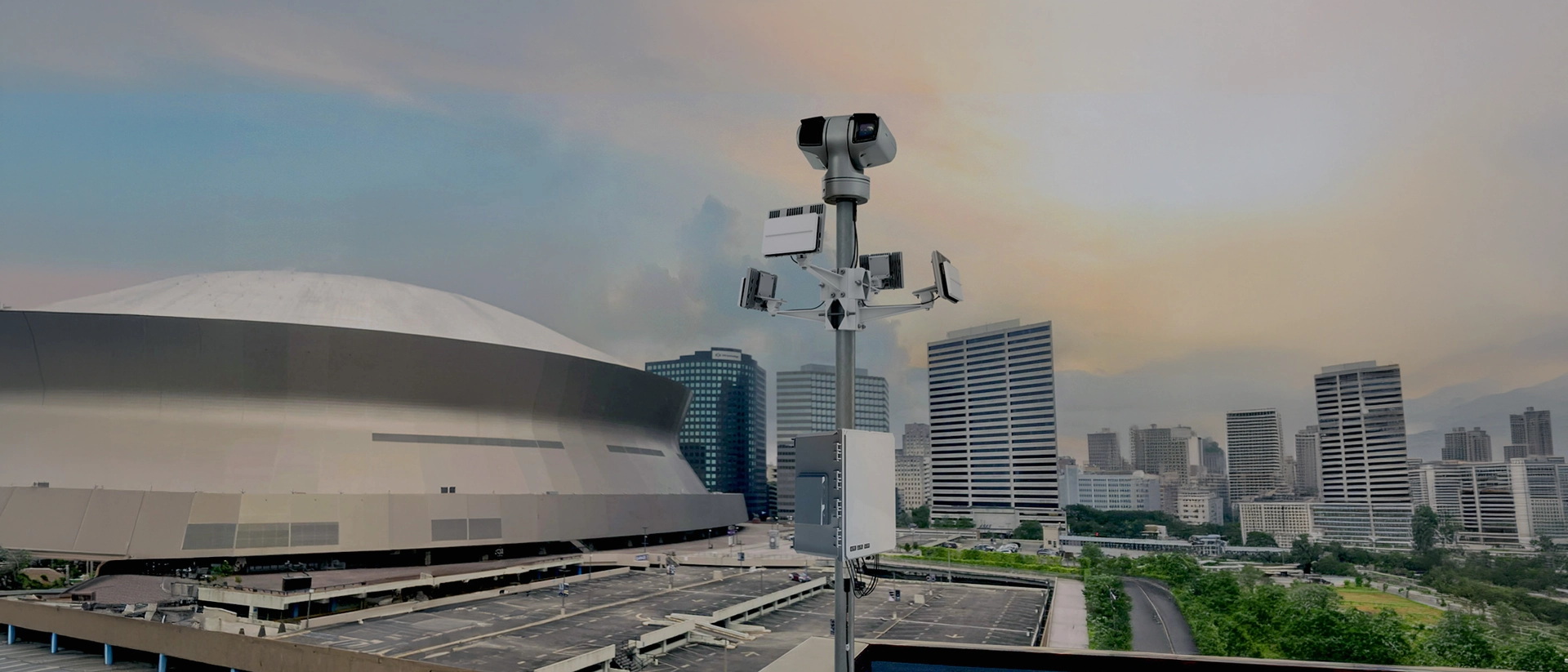 Surveillance camera mounted on pole overlooking a large building and city skyline at sunset.