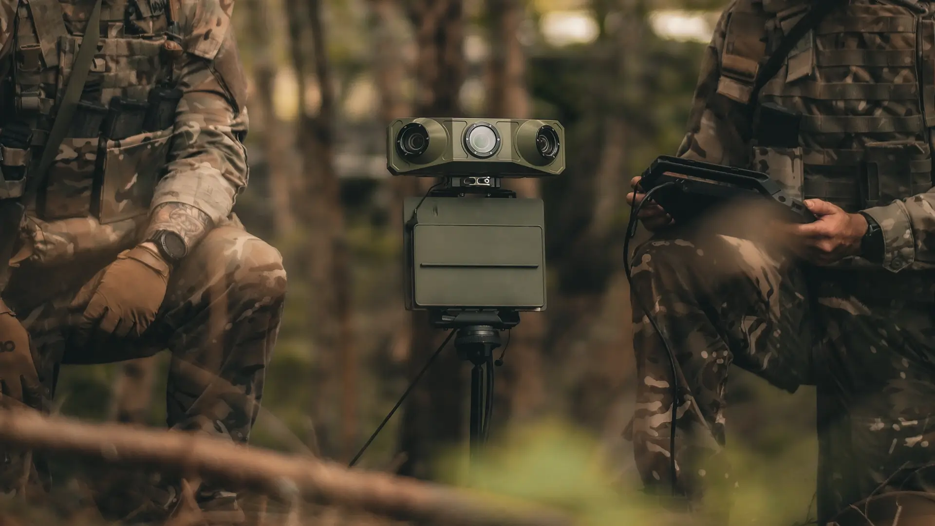 Two soldiers in camouflage operate a small tripod-mounted surveillance robot with cameras in a forest.